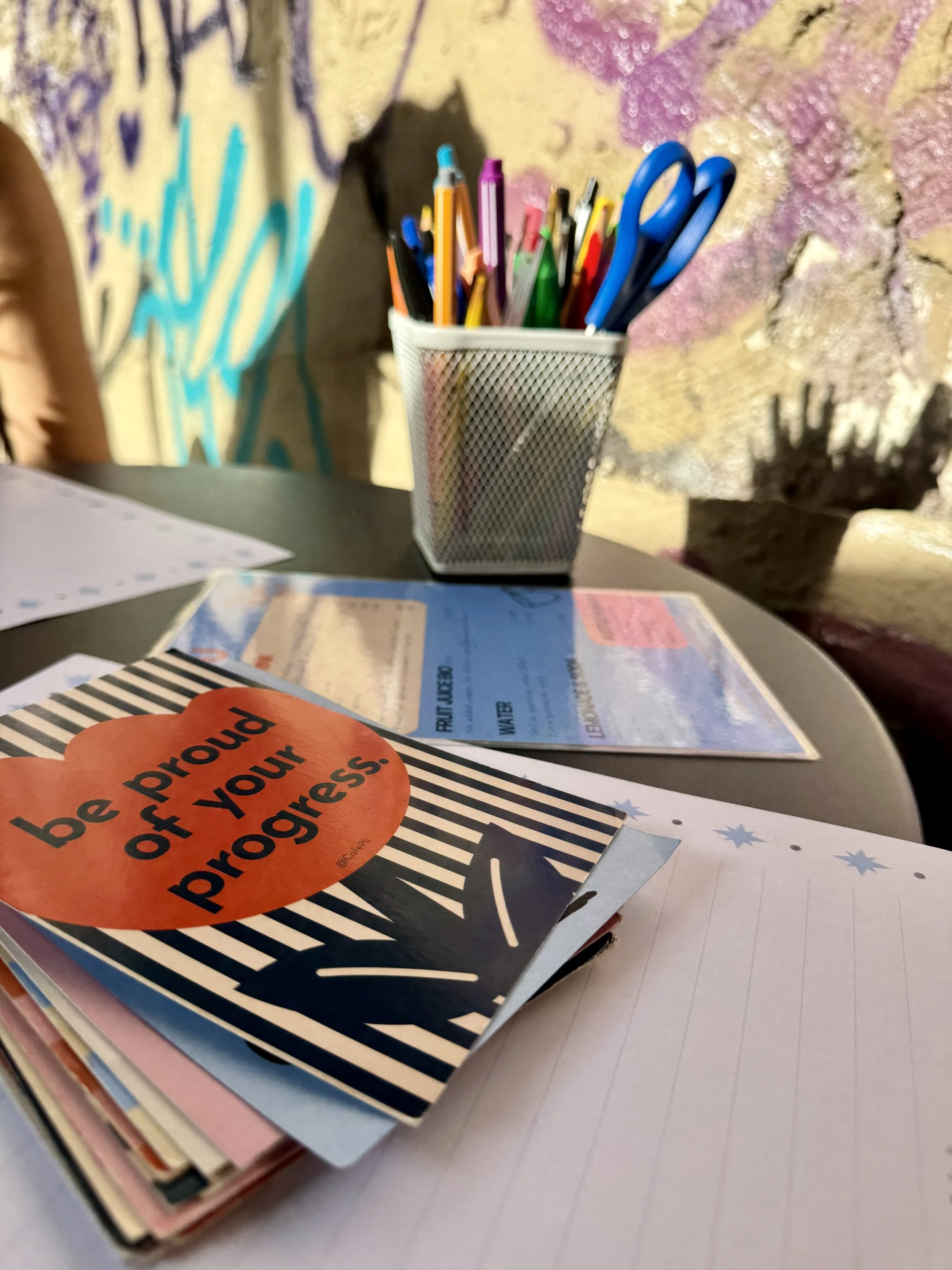 A bright craft desk with colorful pencils and scissors, affirmation cards, and a lined notebook; warm sunlight highlights a message to be proud of your progress.