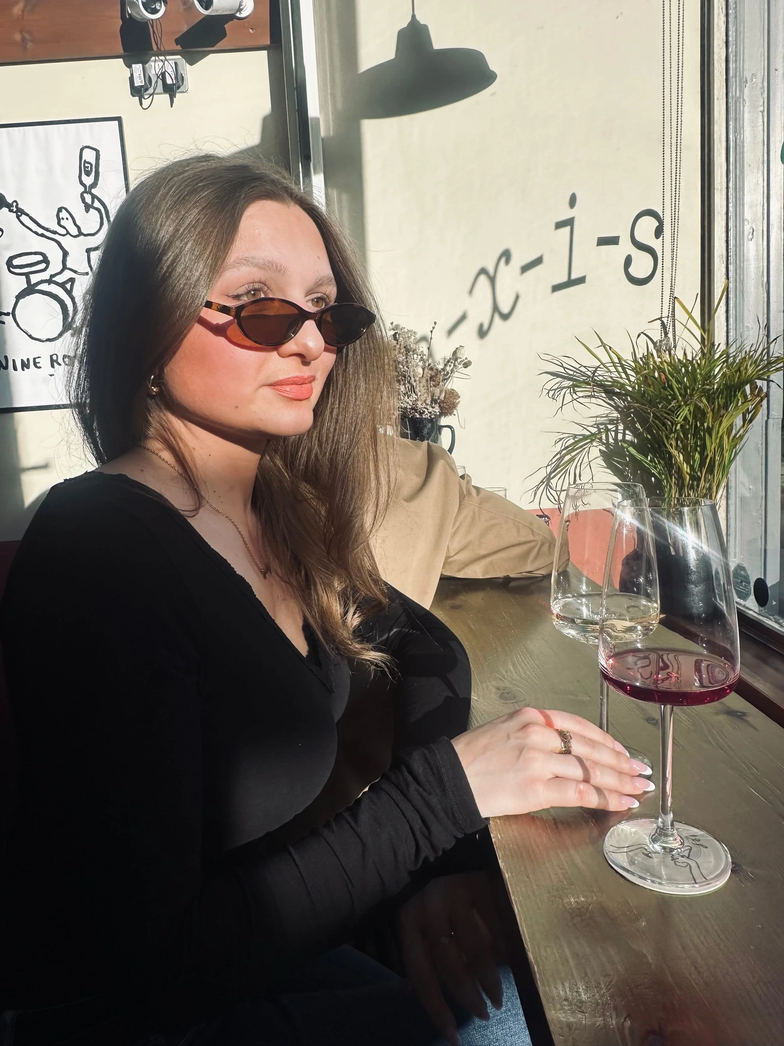 Woman with long hair in sunglasses sits at a sunlit cafe, holding a wine glass. She wears a black top, jeans, and exudes a relaxed vibe.