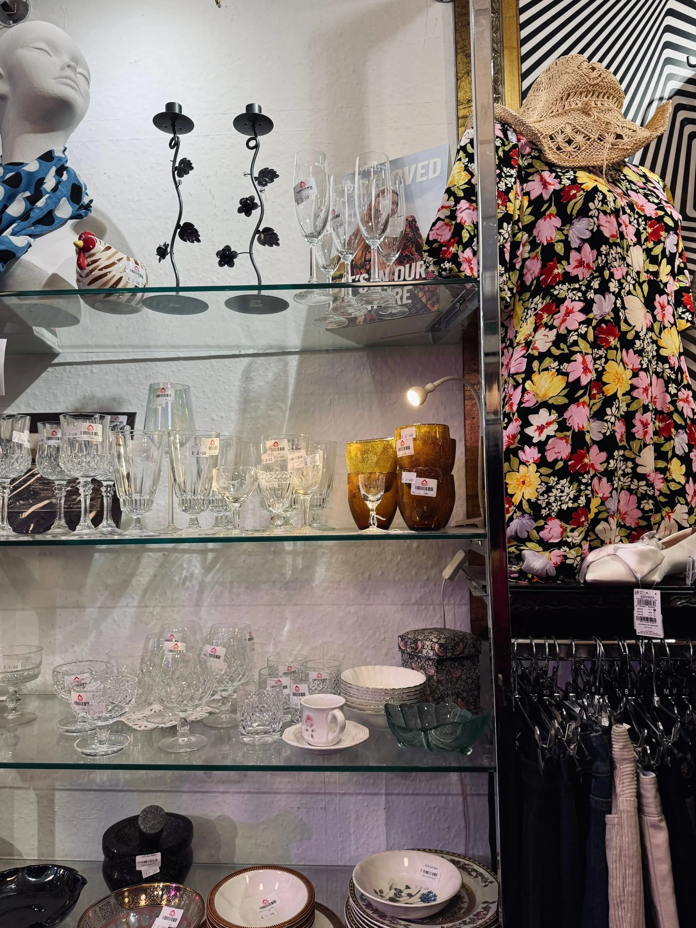 A store display features glass shelves with crystal glassware, patterned bowls, and a mannequin wearing a floral dress and straw hat, creating a vibrant and eclectic setting.