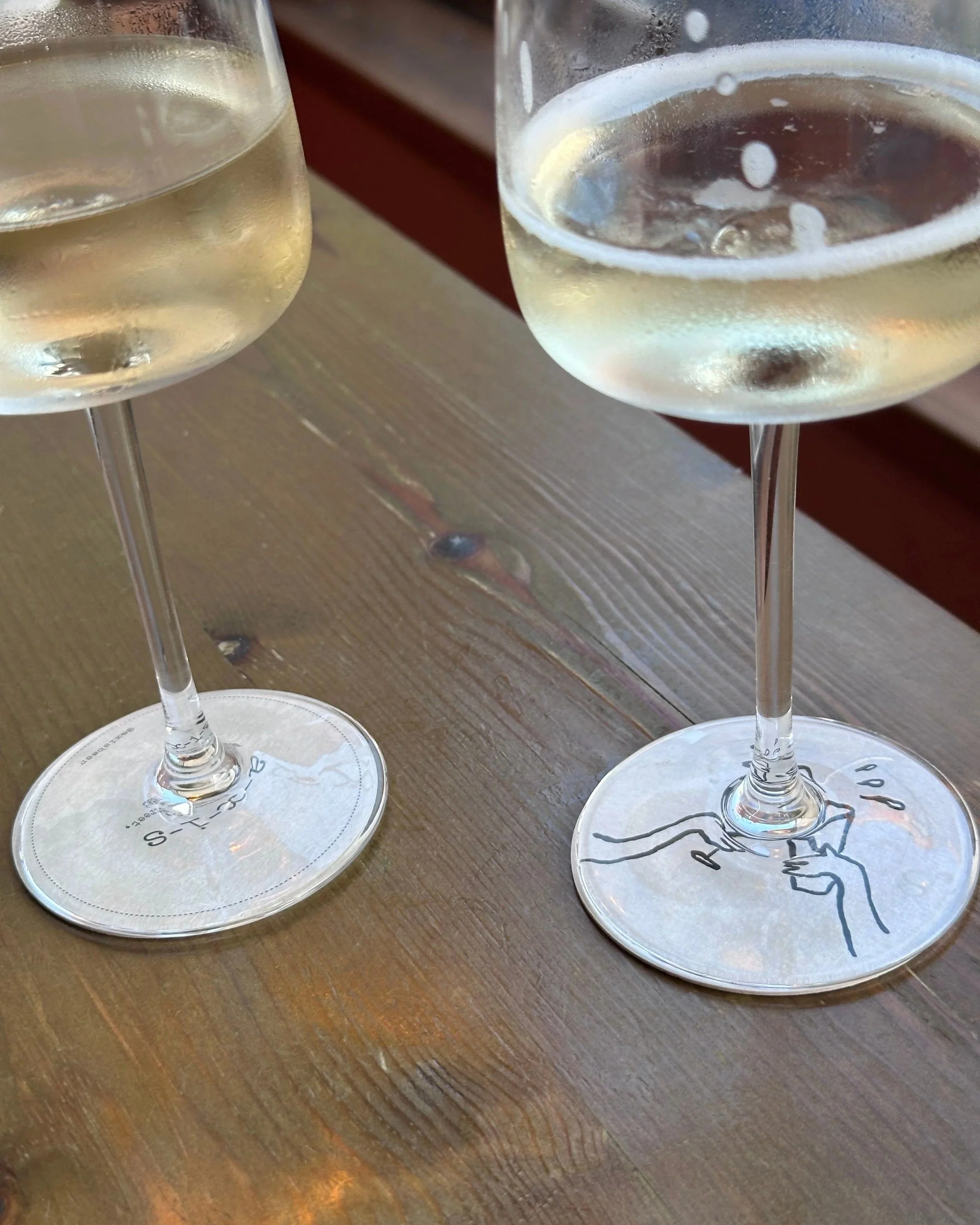 Two wine glasses filled with white wine sit on a wooden table. The glass bases rest on coasters featuring abstract drawings, creating a relaxed, artistic vibe.