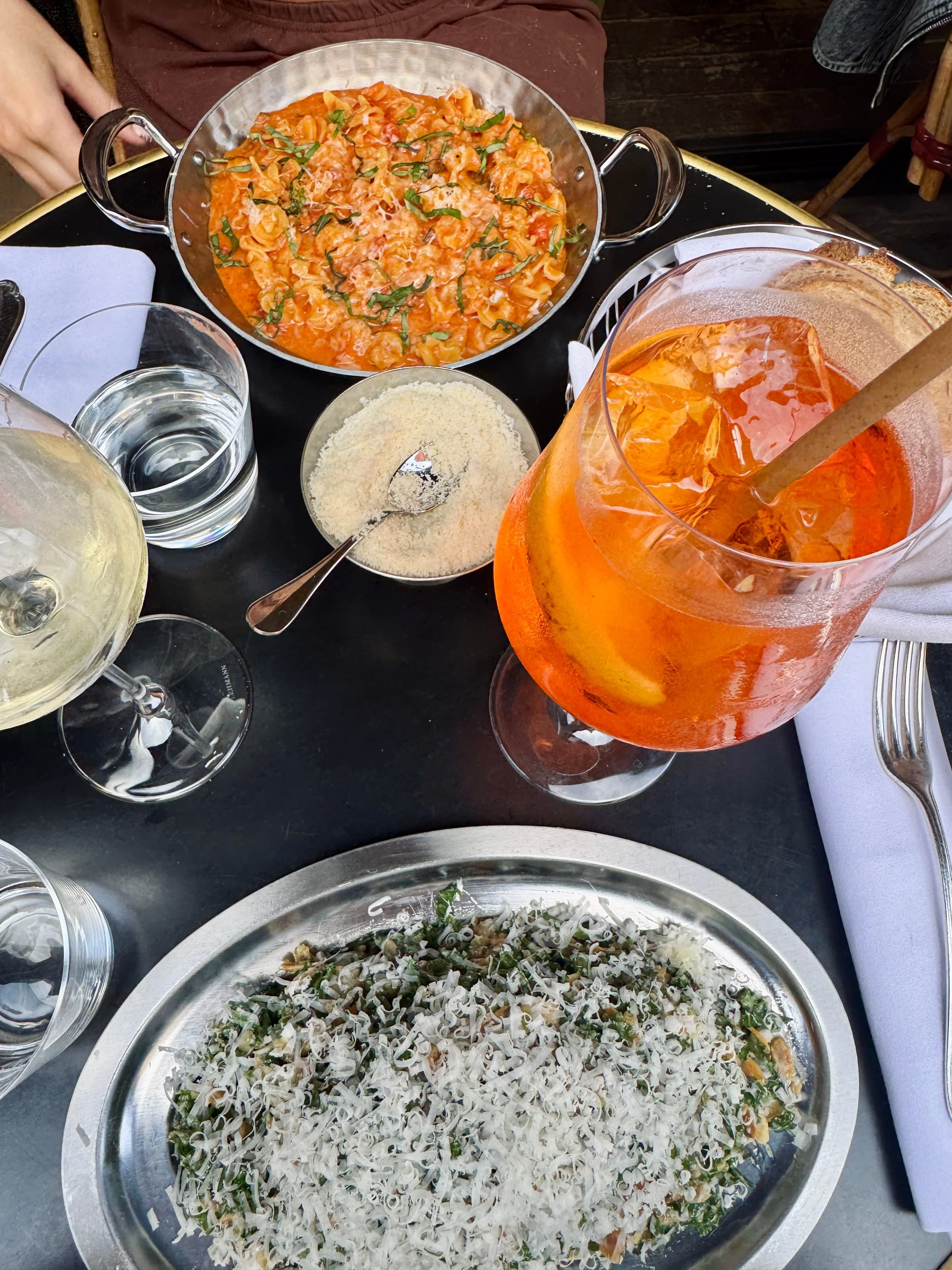 A restaurant table holds tequila pasta, kale salad, bread, drinks, and a small bowl of parmesan; the meal feels lively and inviting.