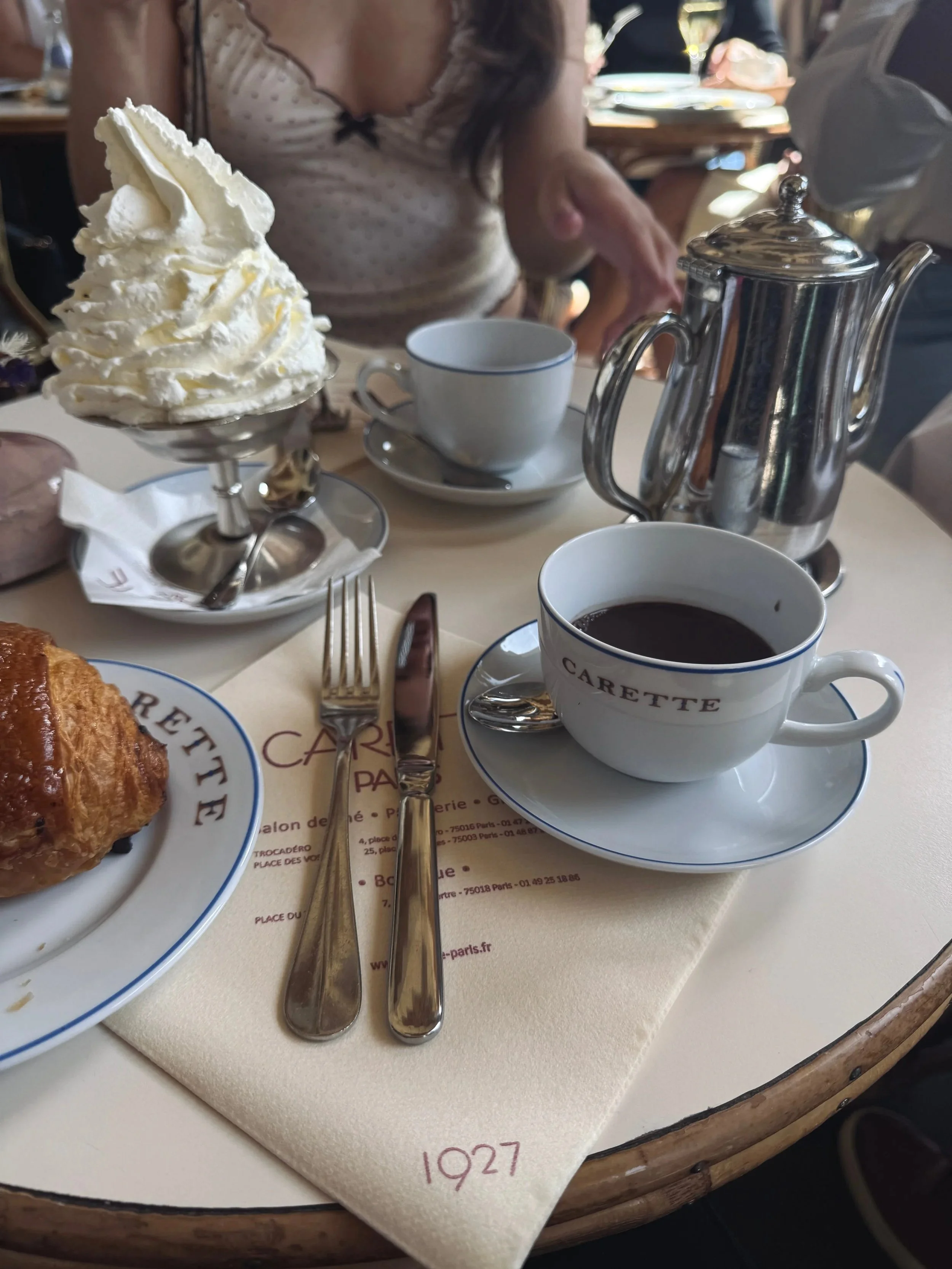 A café table holds espresso in Carette cups, a polished silver teapot, croissant, and tall whipped-cream dessert, creating an elegant Parisian mood.