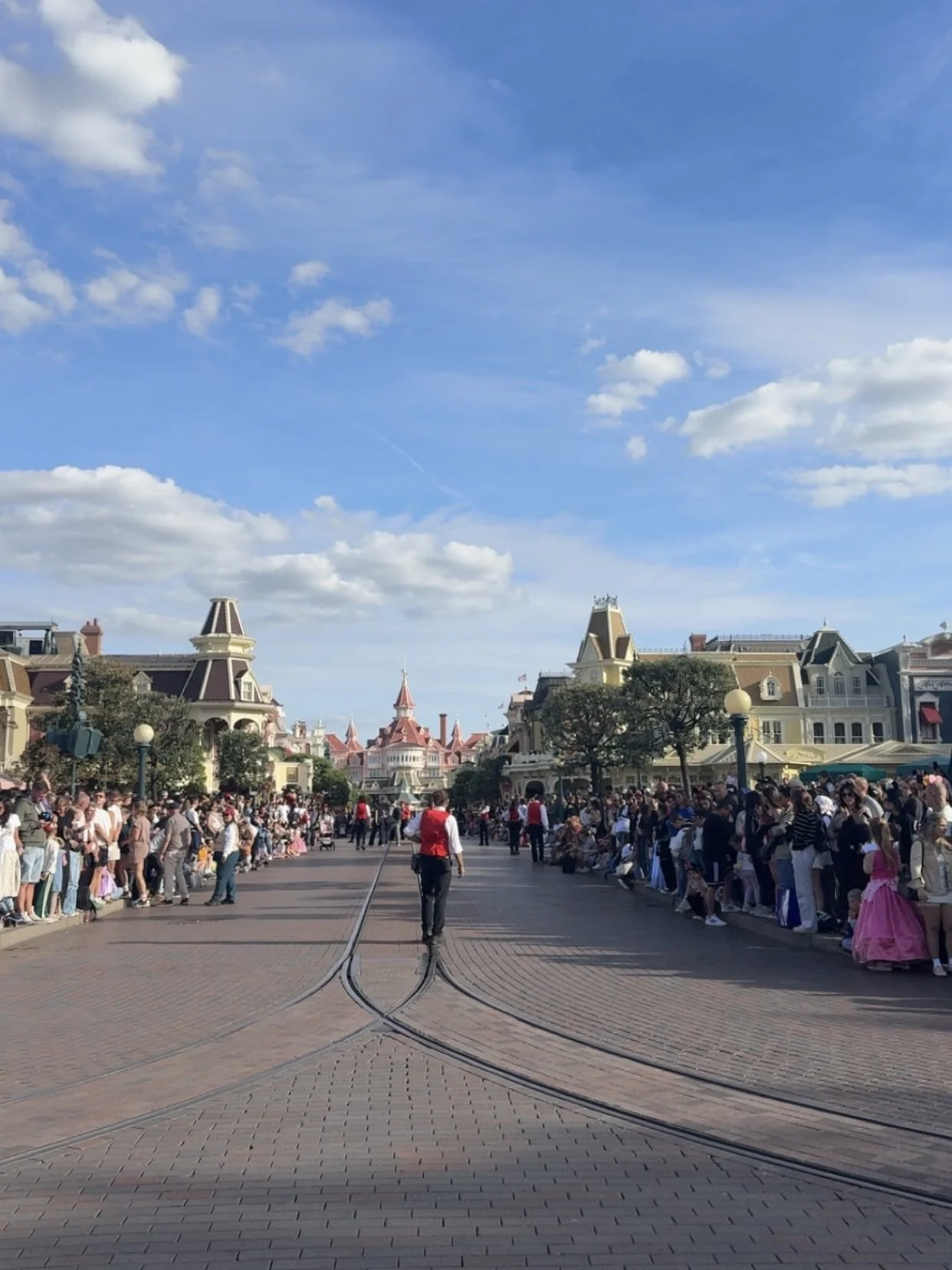 A crowded theme park avenue leads toward a pink fairytale castle beneath a bright blue, cloud-dotted sky; guests line the street, creating a festive mood.
