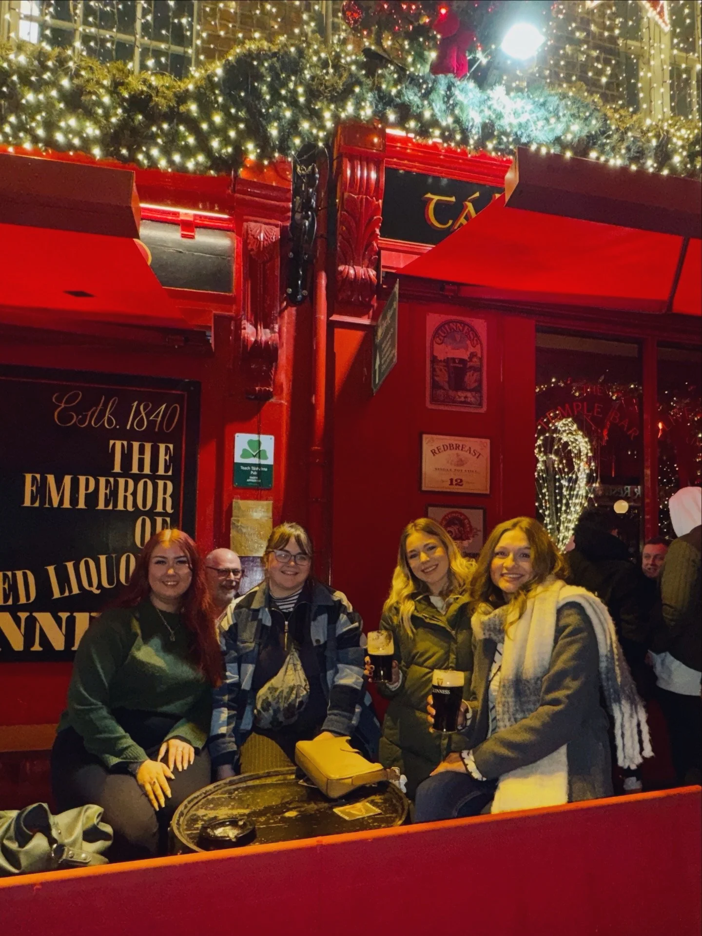 Four girls posing for a photo, sitting outside the Temple Bar in Dublin with pints of Guinness