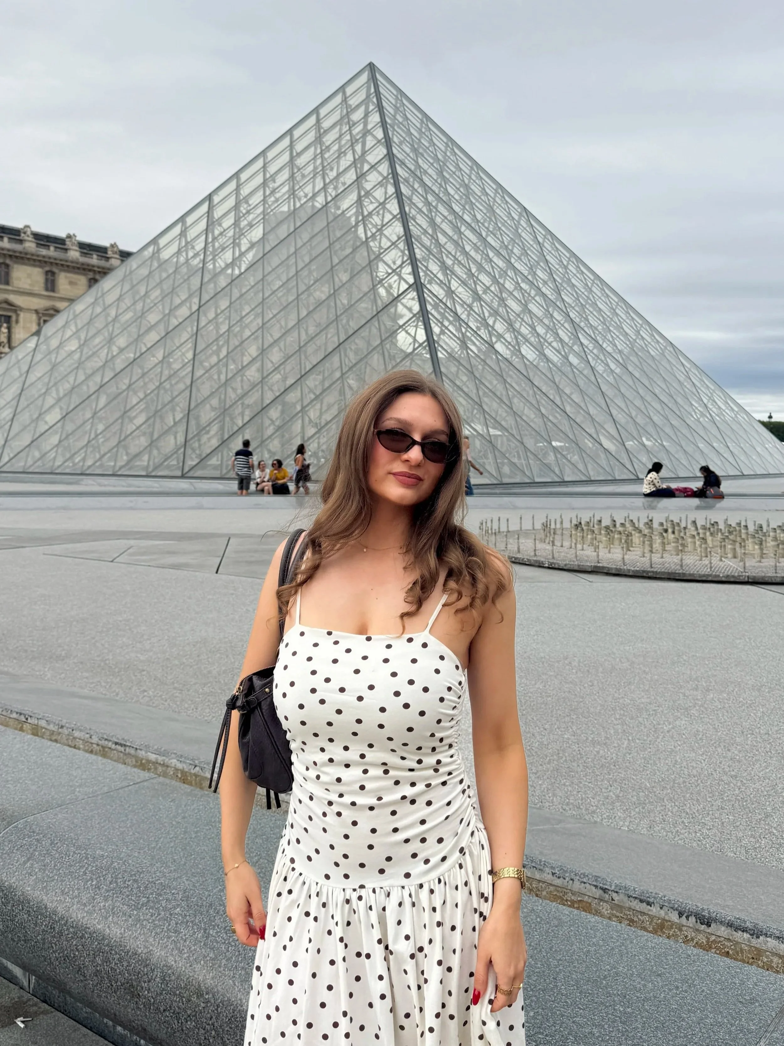 A woman in sunglasses and a white polka-dot dress poses before the Louvre Pyramid in Paris, under a cloudy sky, in a relaxed travel snapshot.