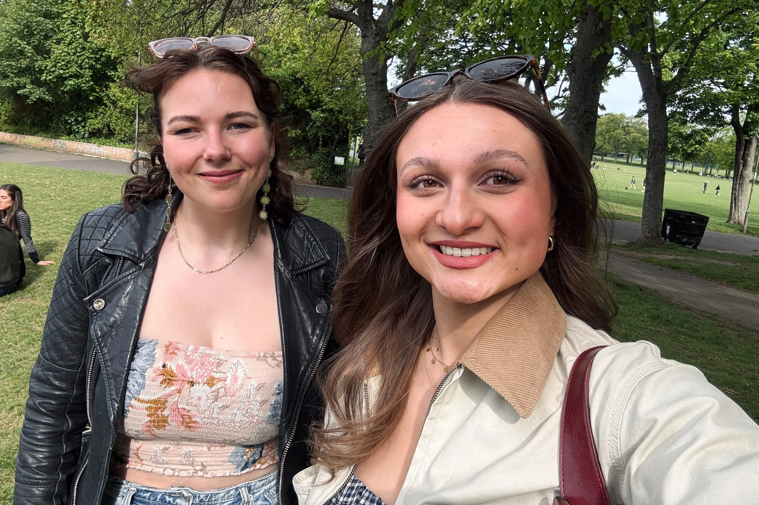 Two women smiling in a sunny park. One wears a black leather jacket, the other a beige coat. Trees and people sitting on grass in the background.