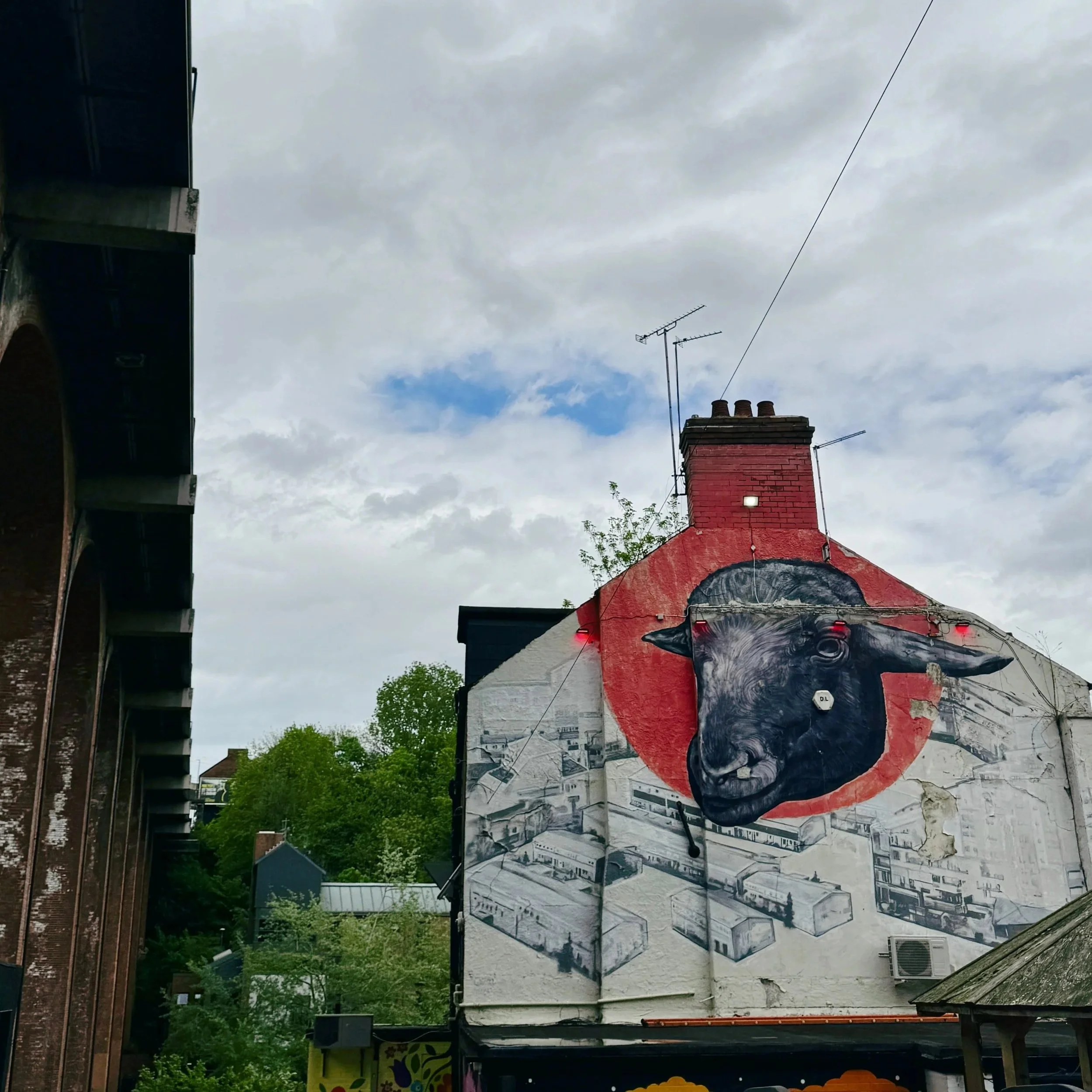 Street art on a building depicts a large black cow's head in front of a red circle. A bridge and cloudy sky fill the background, creating an urban feel.