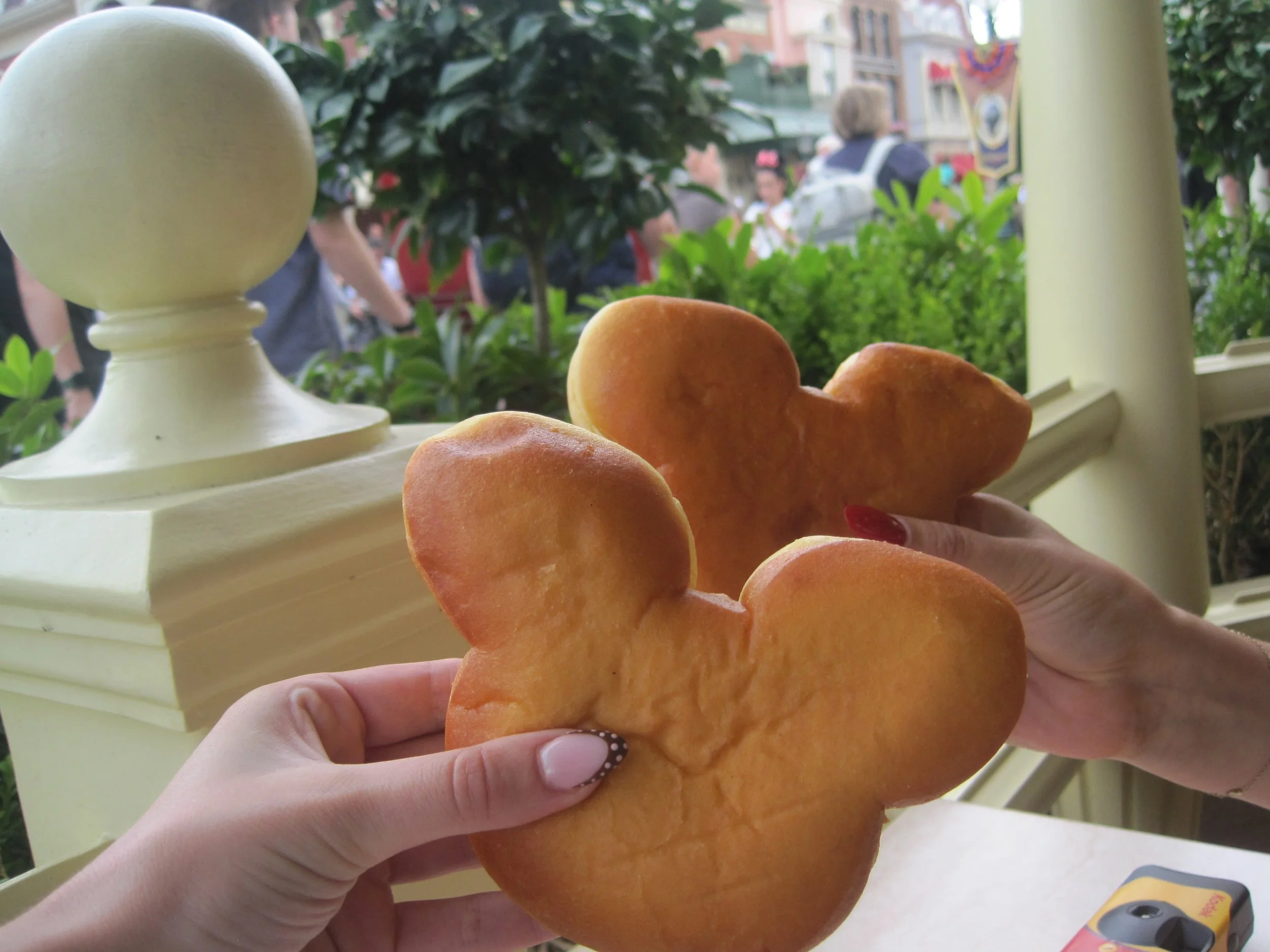 Hands hold two golden-brown Mickey Mouse-shaped pastries by a café window, with leafy plants and a lively street outside; a cheerful, playful scene.