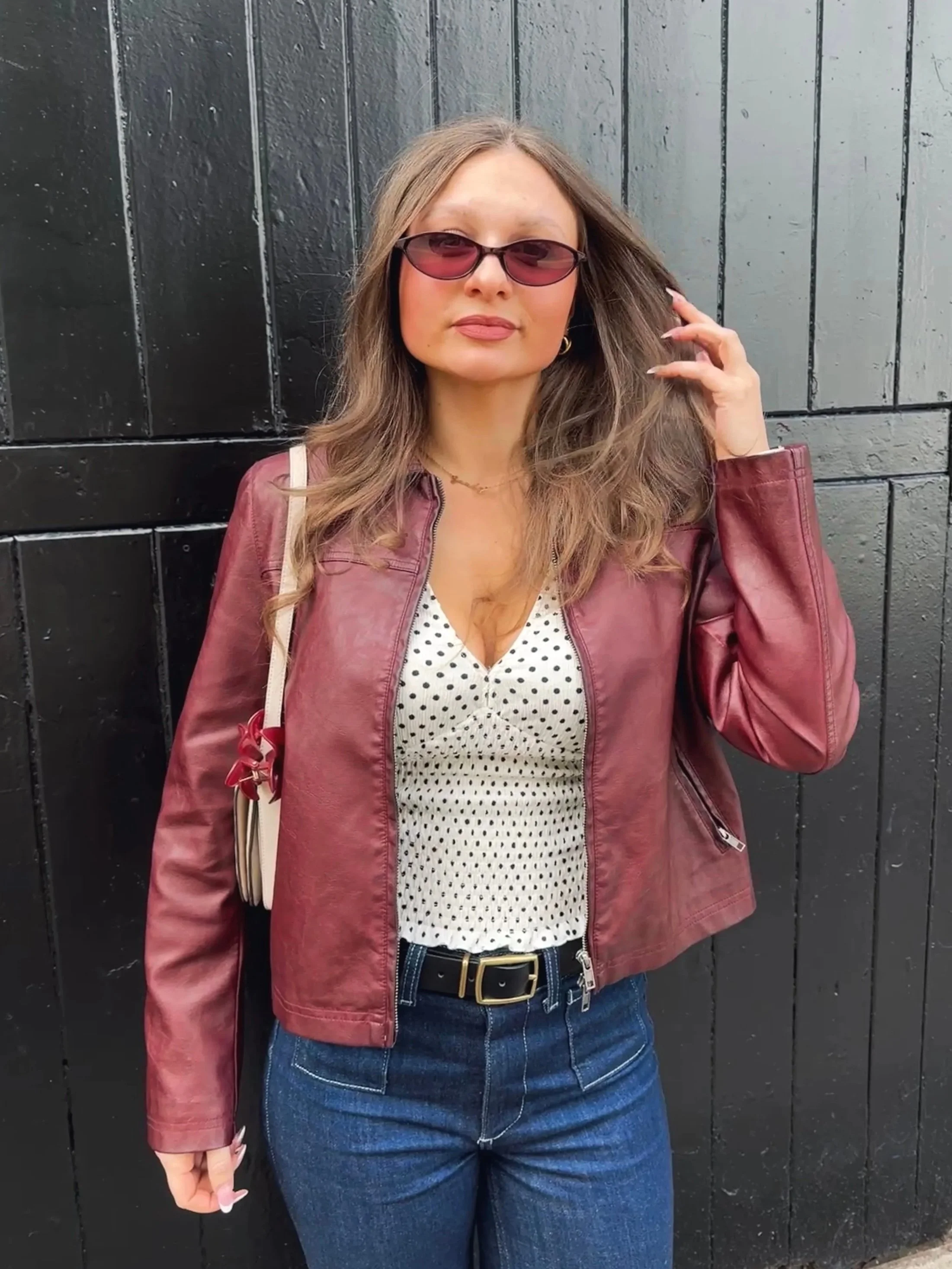 Woman in sunglasses, maroon jacket, and polka-dot top stands confidently against a black wooden background.