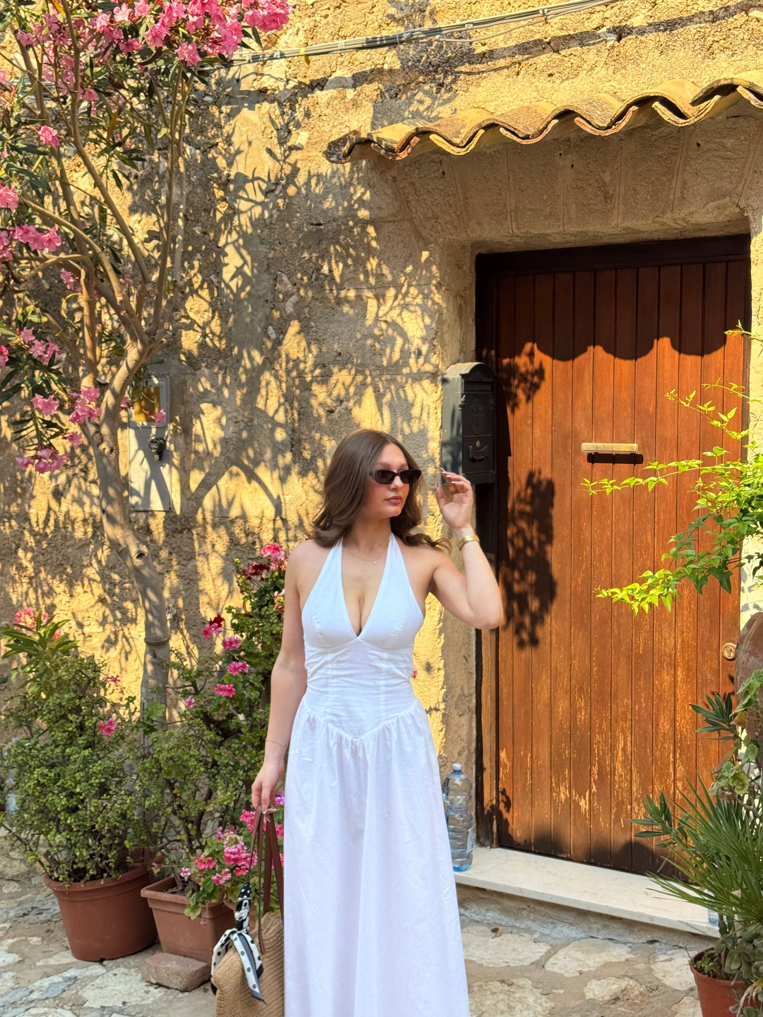A woman wearing a white dress poses in front of a cozy house, showcasing a serene and inviting atmosphere.
