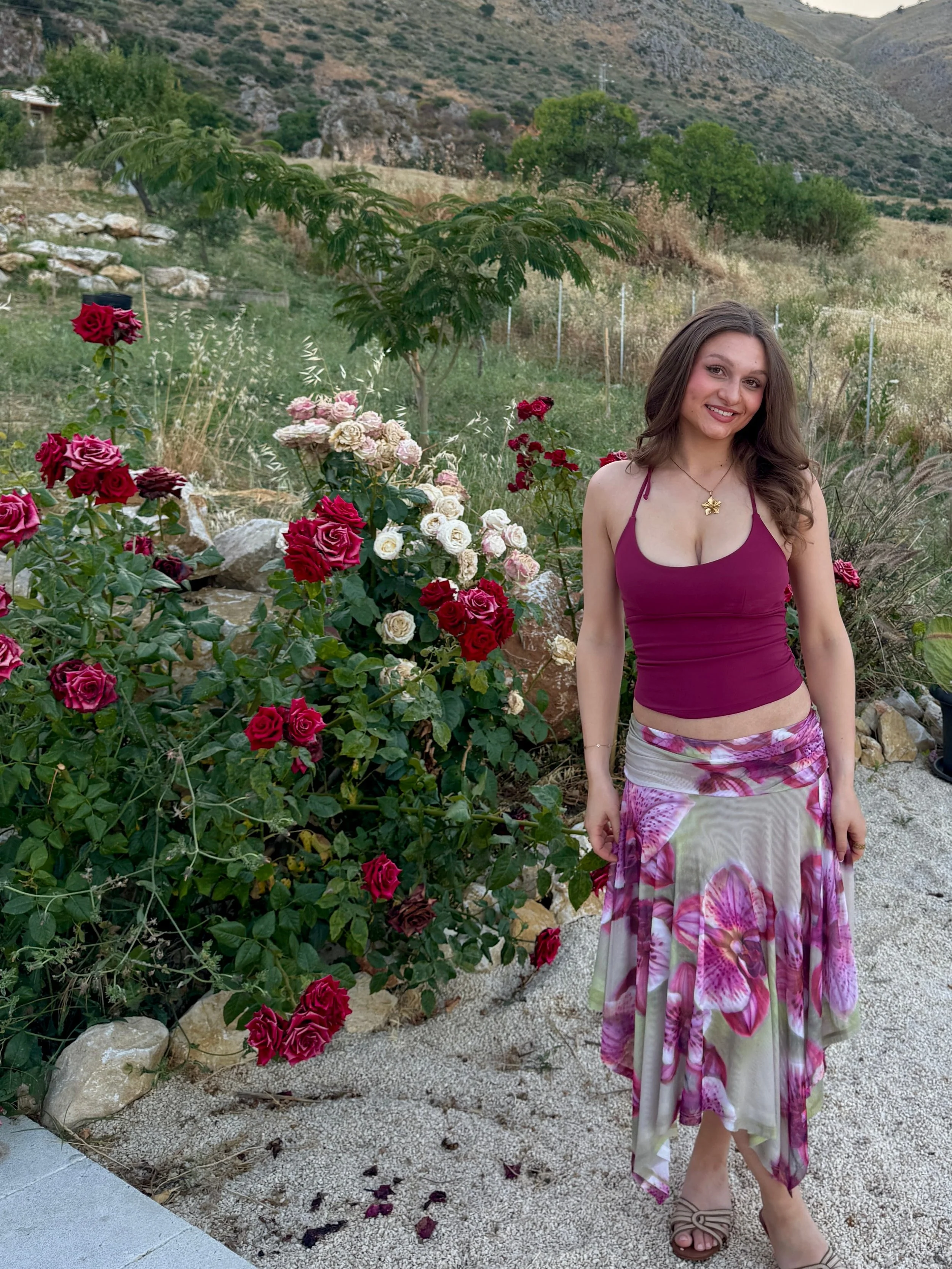 A woman smiles while standing in front of a vibrant flower garden, surrounded by colorful blooms.