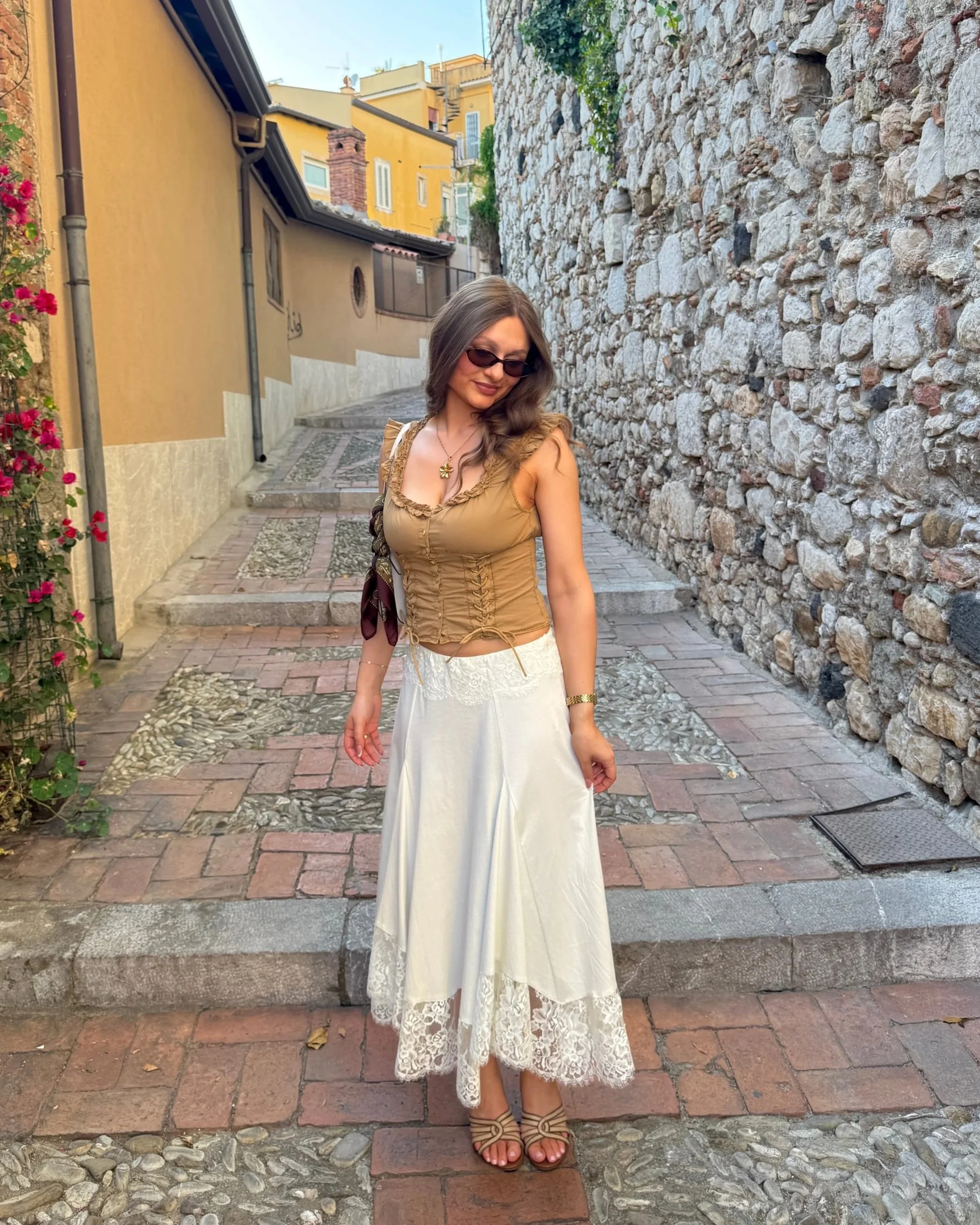 Woman in sunglasses wearing a tan top and white skirt stands on a cobblestone path beside a stone wall and flowering vine.