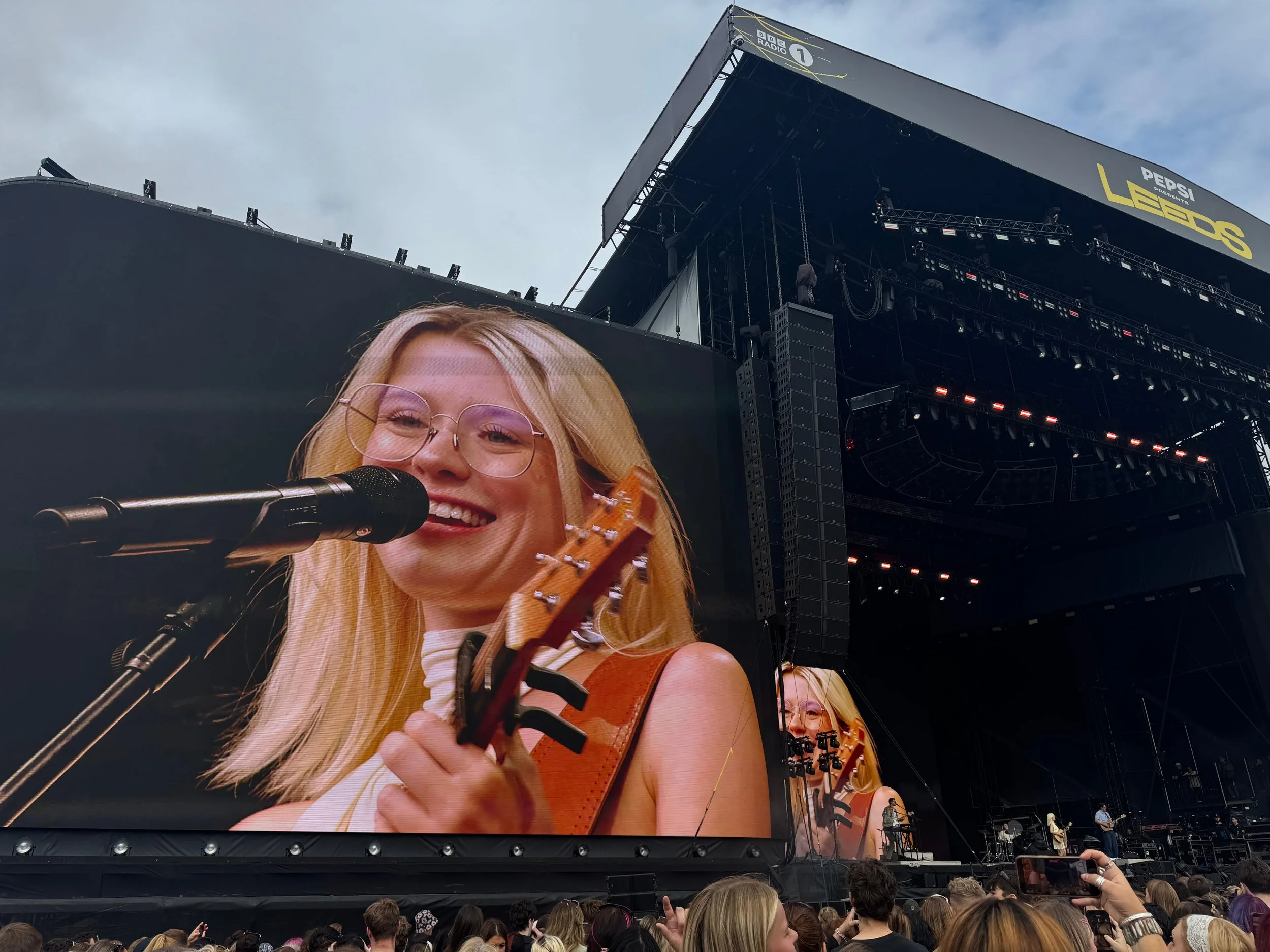 Singer Maisie Peters sings into a microphone on a large festival screen while performing on a dark outdoor stage before a crowd.