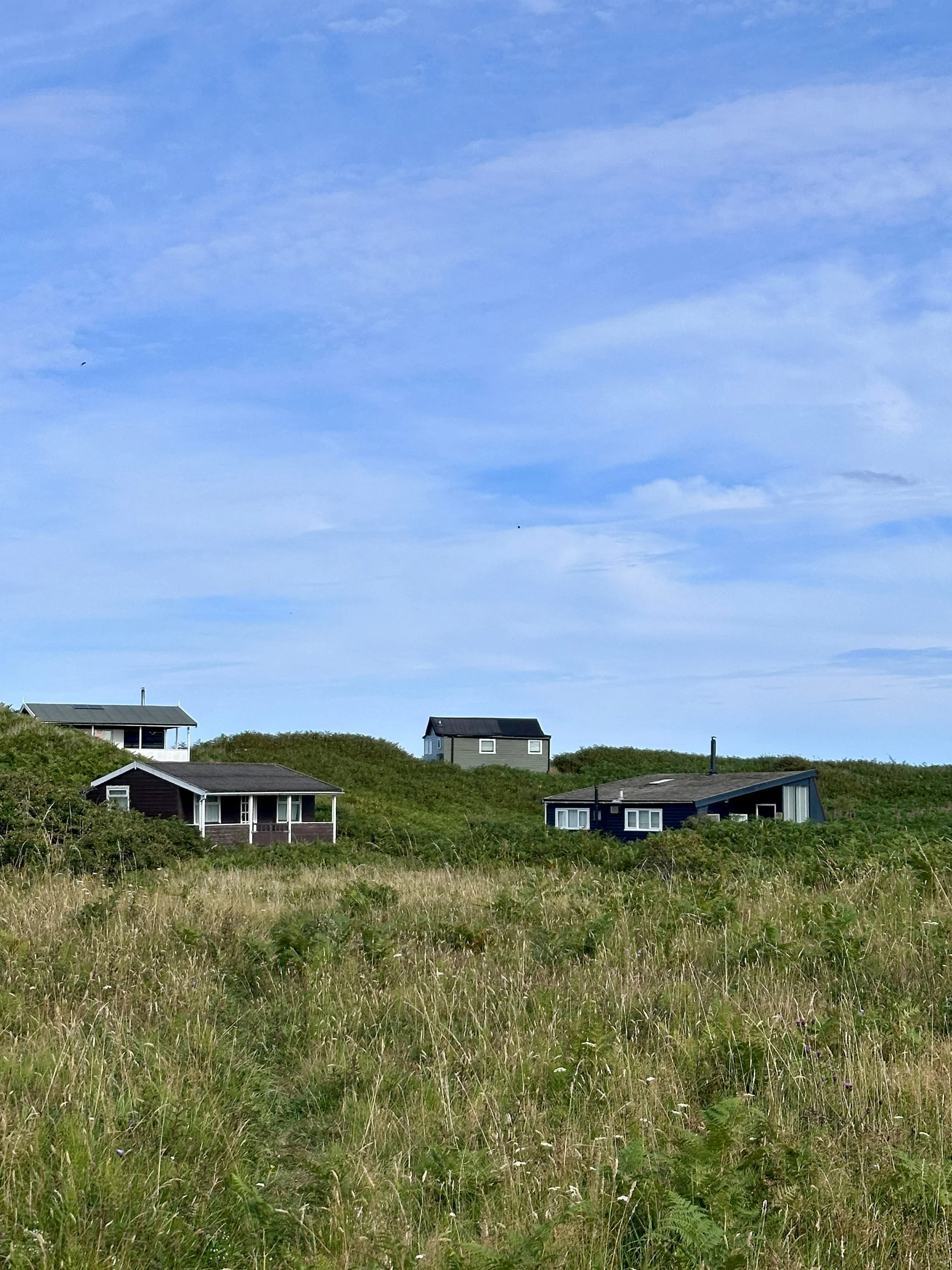 Small coastal cottages dot grassy dunes beneath a vast blue, cloud-streaked sky, creating a quiet, breezy, serene seaside landscape.