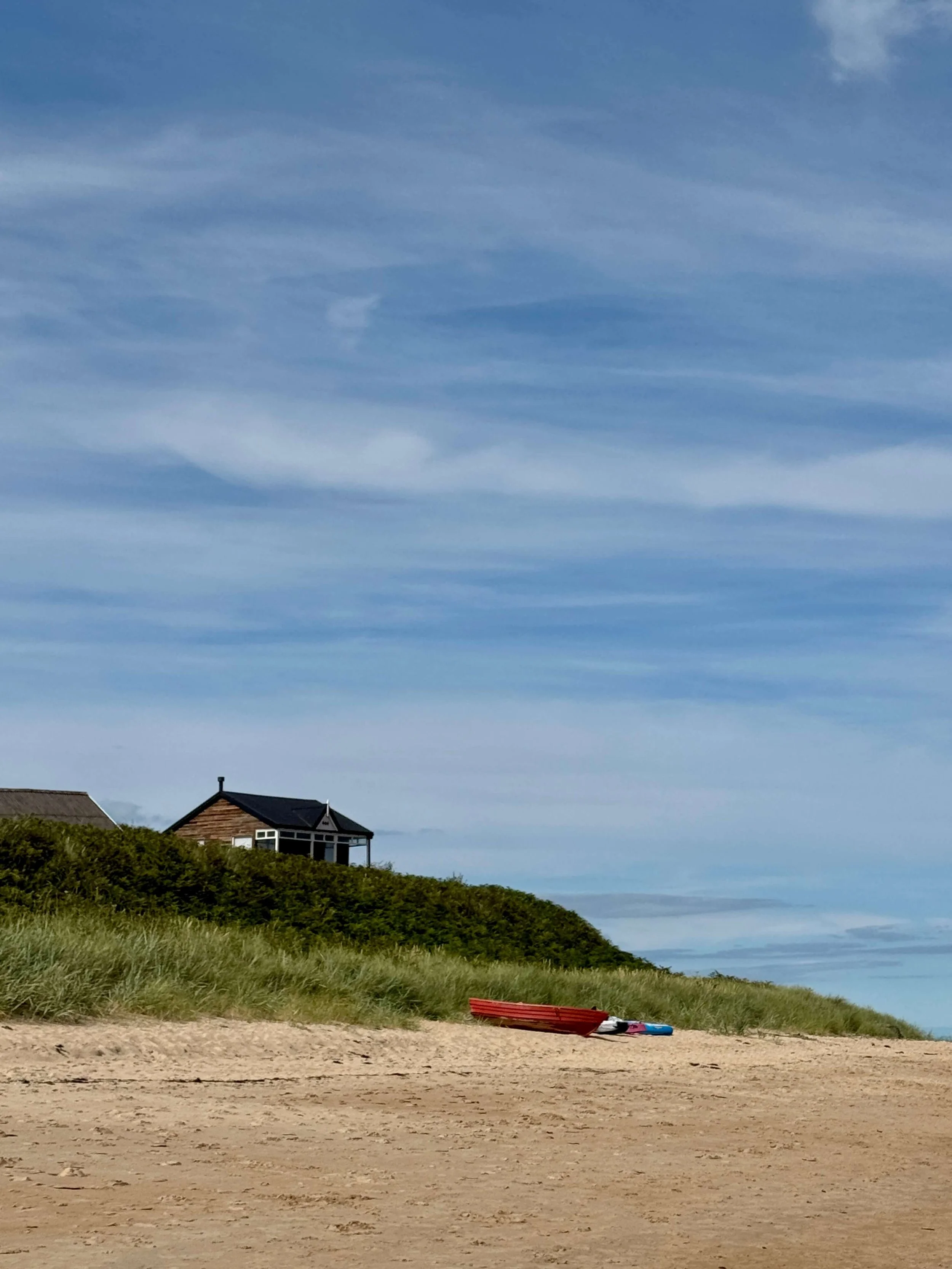 Quiet sandy beach with dune grass, a seaside cottage, and red kayaks beneath a vast blue, cloud-streaked sky; calm coastal mood.
