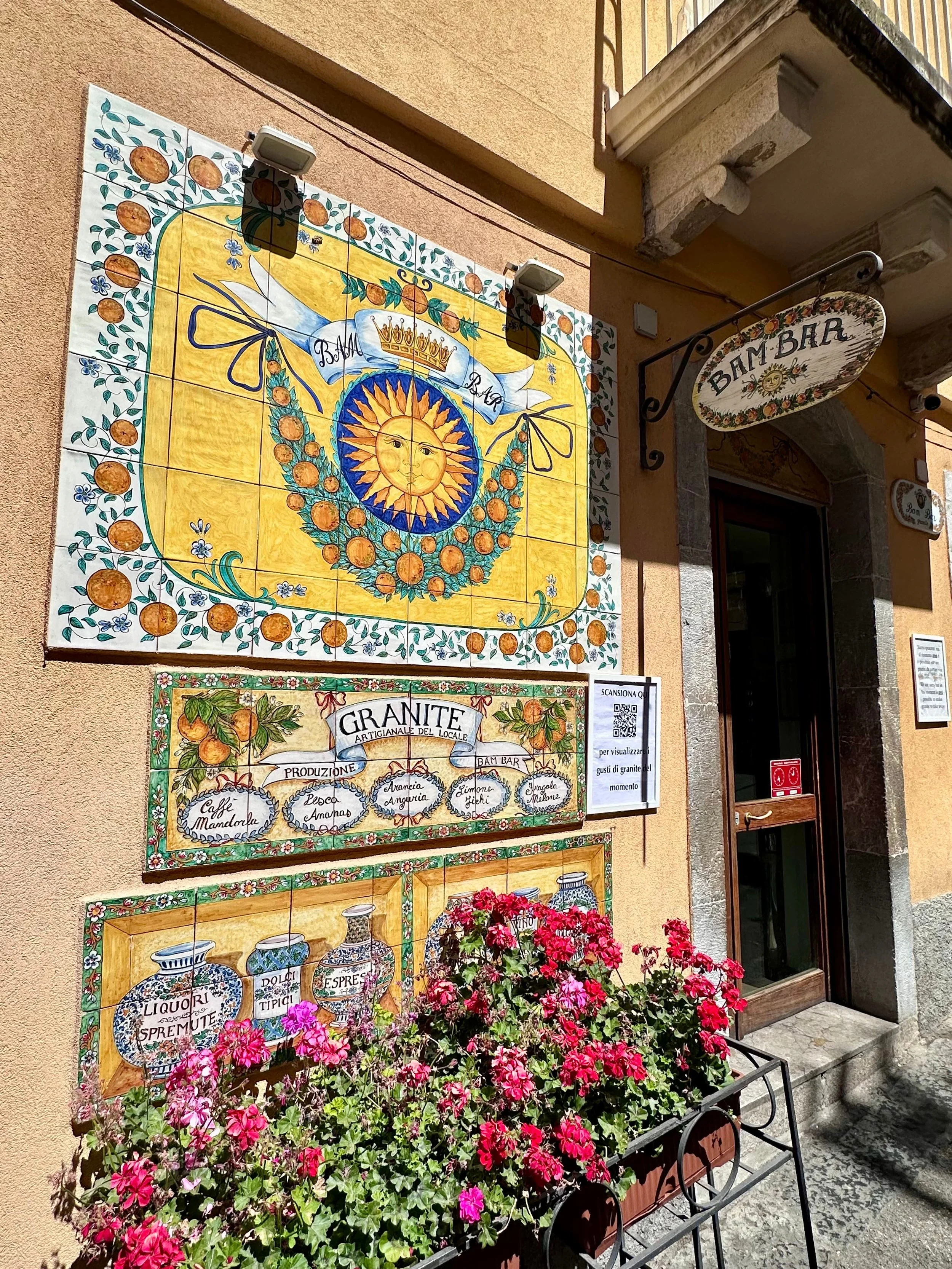 Bright outdoor wall with colorful ceramic tiles featuring sun and fruit designs, a sign reading "Bam Bar," and a planter with vibrant pink flowers.