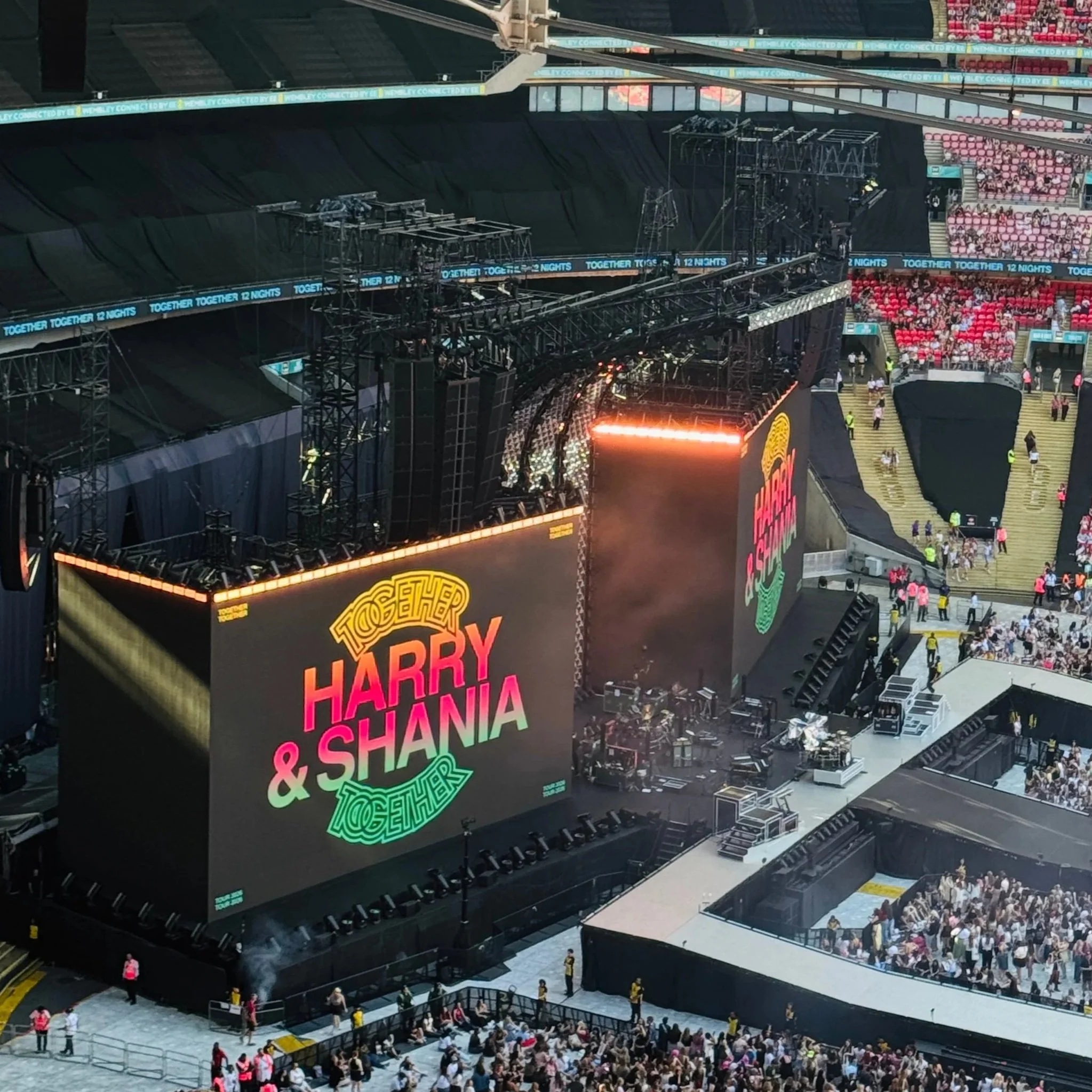 A vibrant crowd at a Harry Styles concert in Wembley, featuring a stage and a large screen displaying visuals
