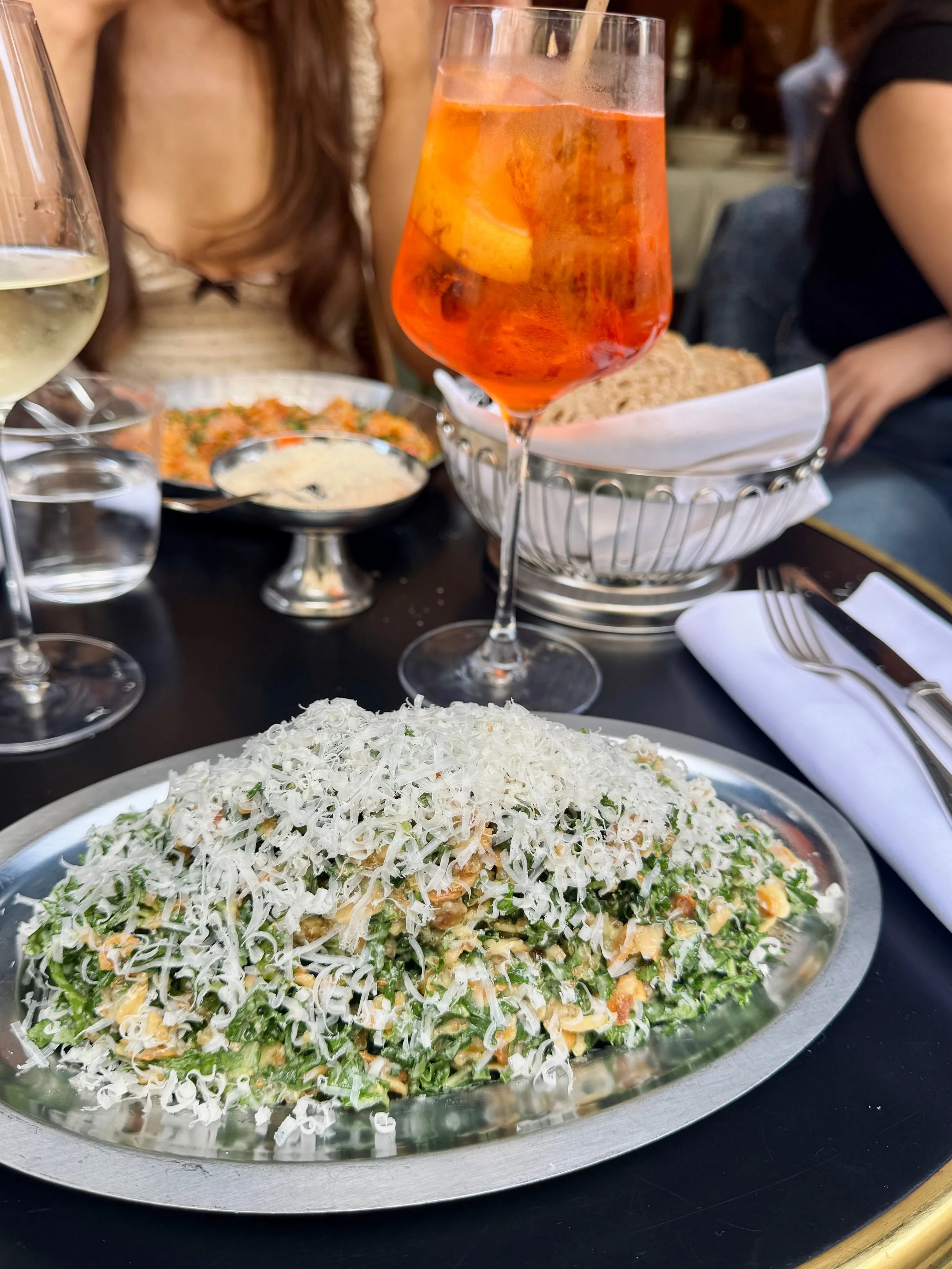 A richly topped pasta dish with greens and grated cheese sits on a restaurant table beside an orange spritz cocktail and wine, creating a lively dining mood.