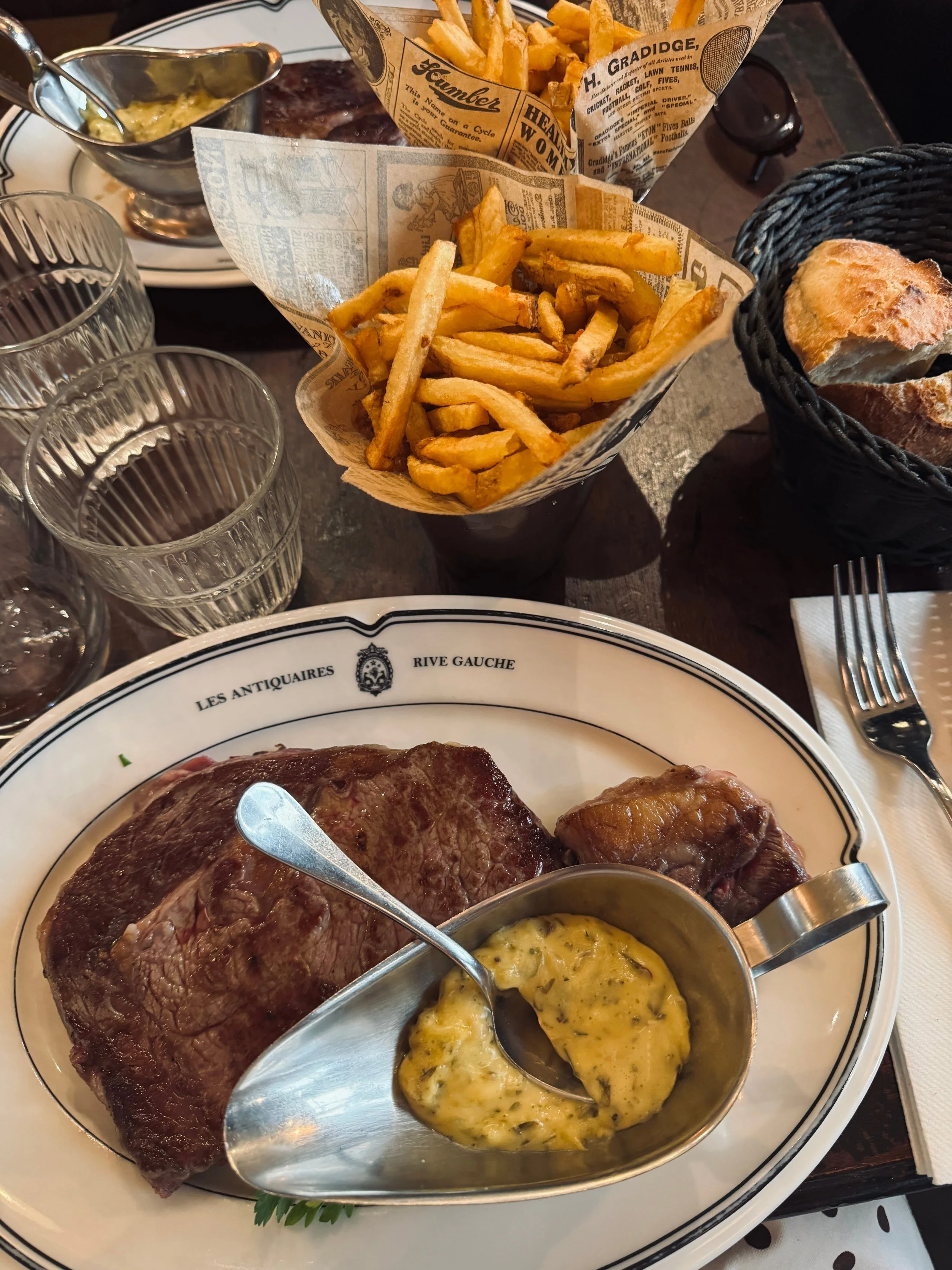 A bistro steak dinner features rib steak, bearnaise sauce, golden fries in newspaper-lined baskets, bread, and water glasses on a dark table.