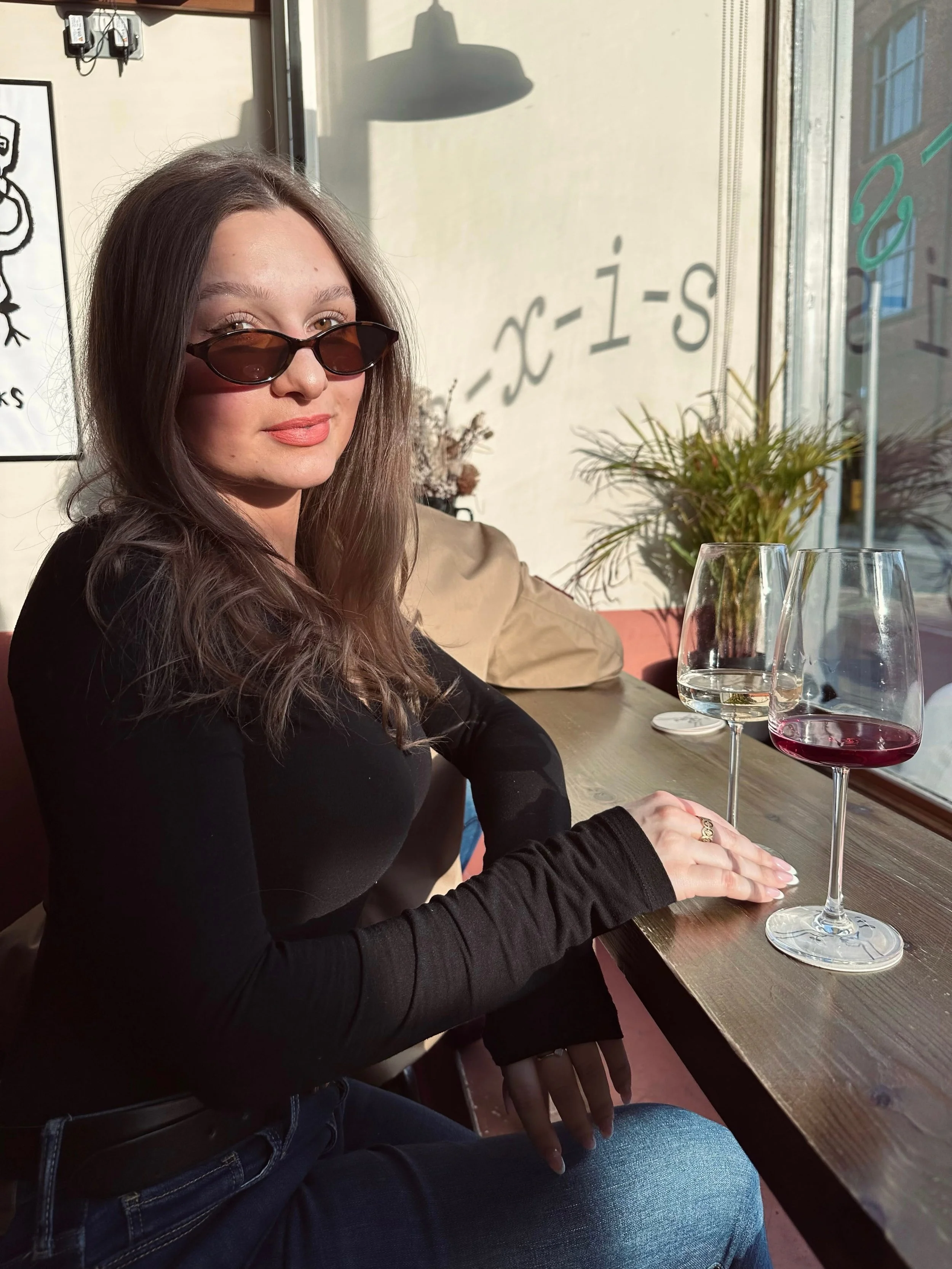 Woman with long hair in sunglasses sits at a sunlit cafe, holding a wine glass. She wears a black top, jeans, and exudes a relaxed vibe.