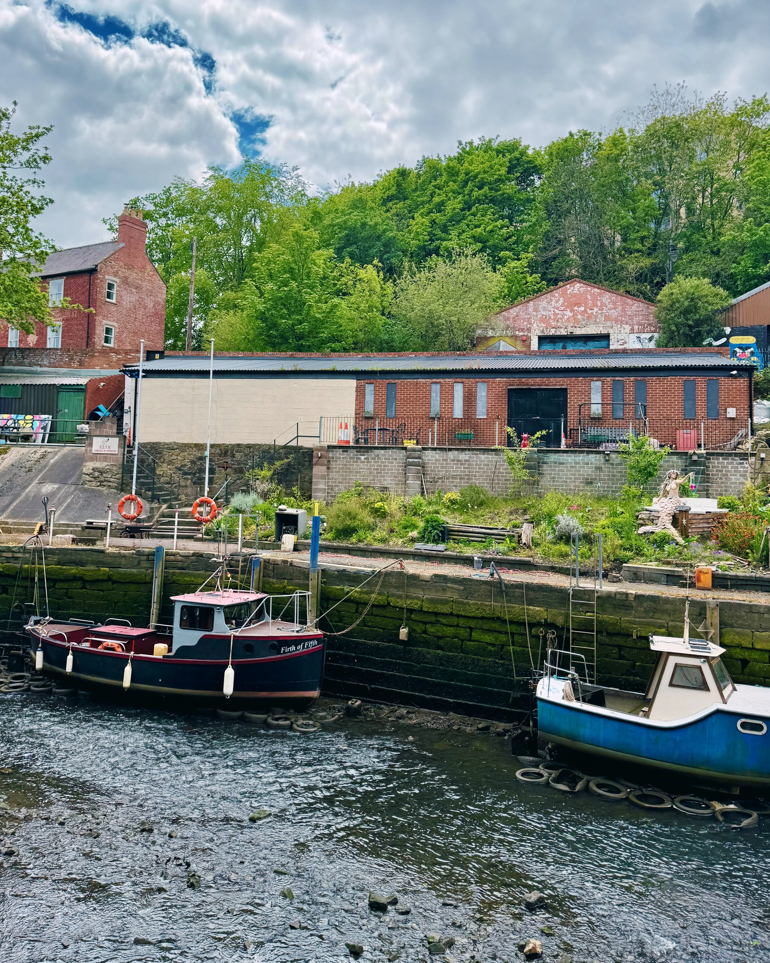 A river with two boats rests at low tide against a stone wall. Behind them, brick buildings and green trees stand under a cloudy sky, creating a serene atmosphere.