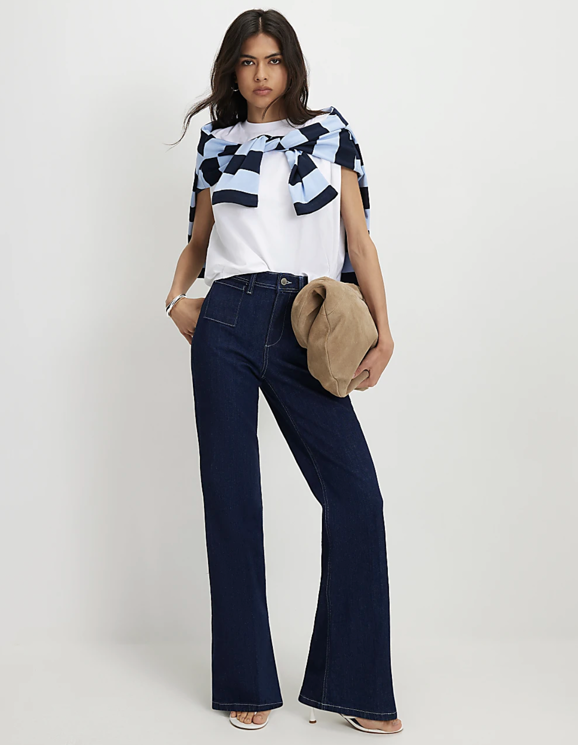 Woman in a white top with a blue checkered sweater draped over shoulders, wearing dark flare jeans and holding a tan purse.
