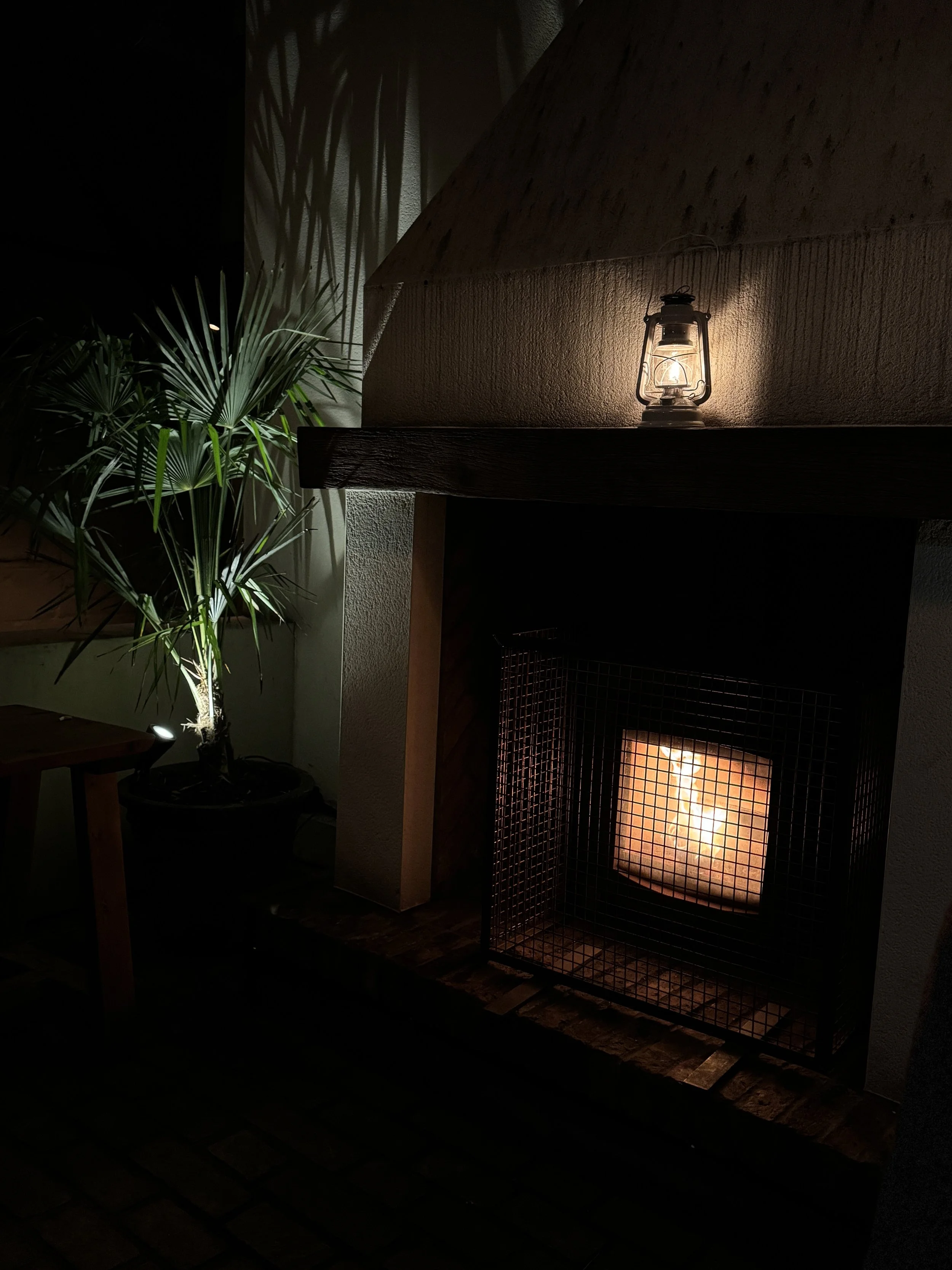 A glowing fireplace with a lantern sitting on the mantle piece, and a potted plant in the background
