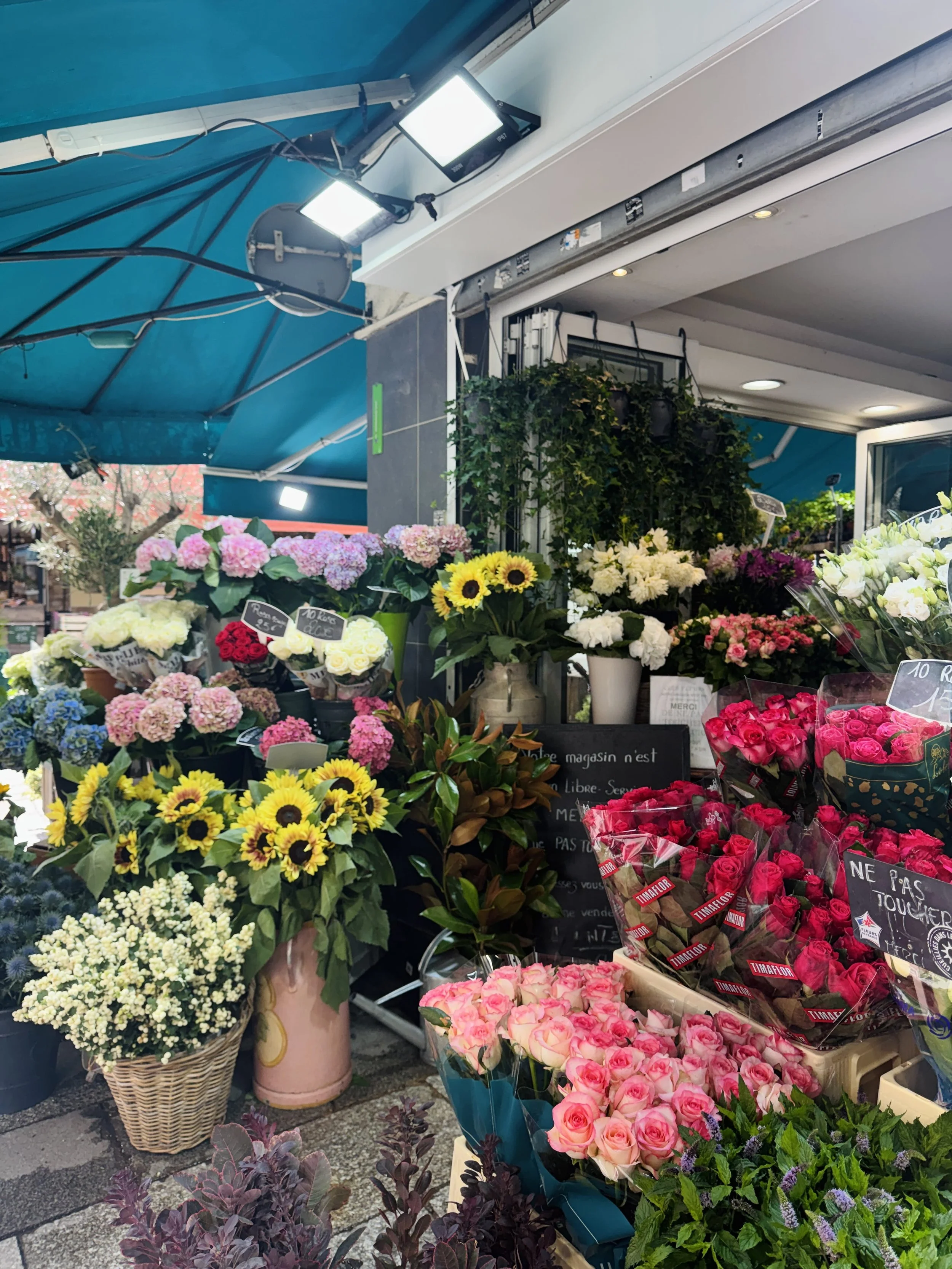 A vibrant flower shop under a teal awning displays colorful bouquets of roses, sunflowers, hydrangeas, and lilies, creating a lively, welcoming street-market scene.