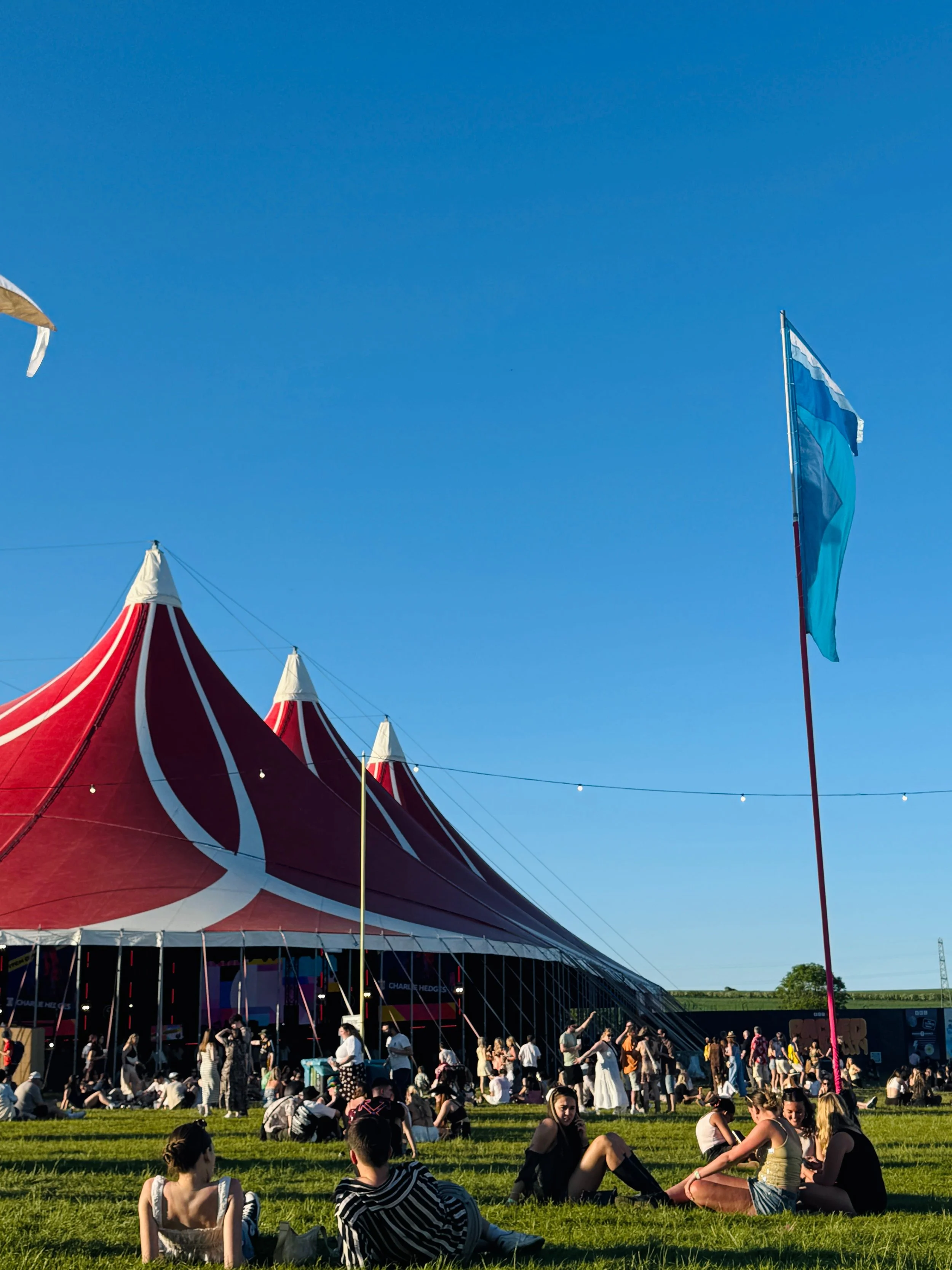 Festival attendees relax on grass near a large red and white striped tent under a clear blue sky.