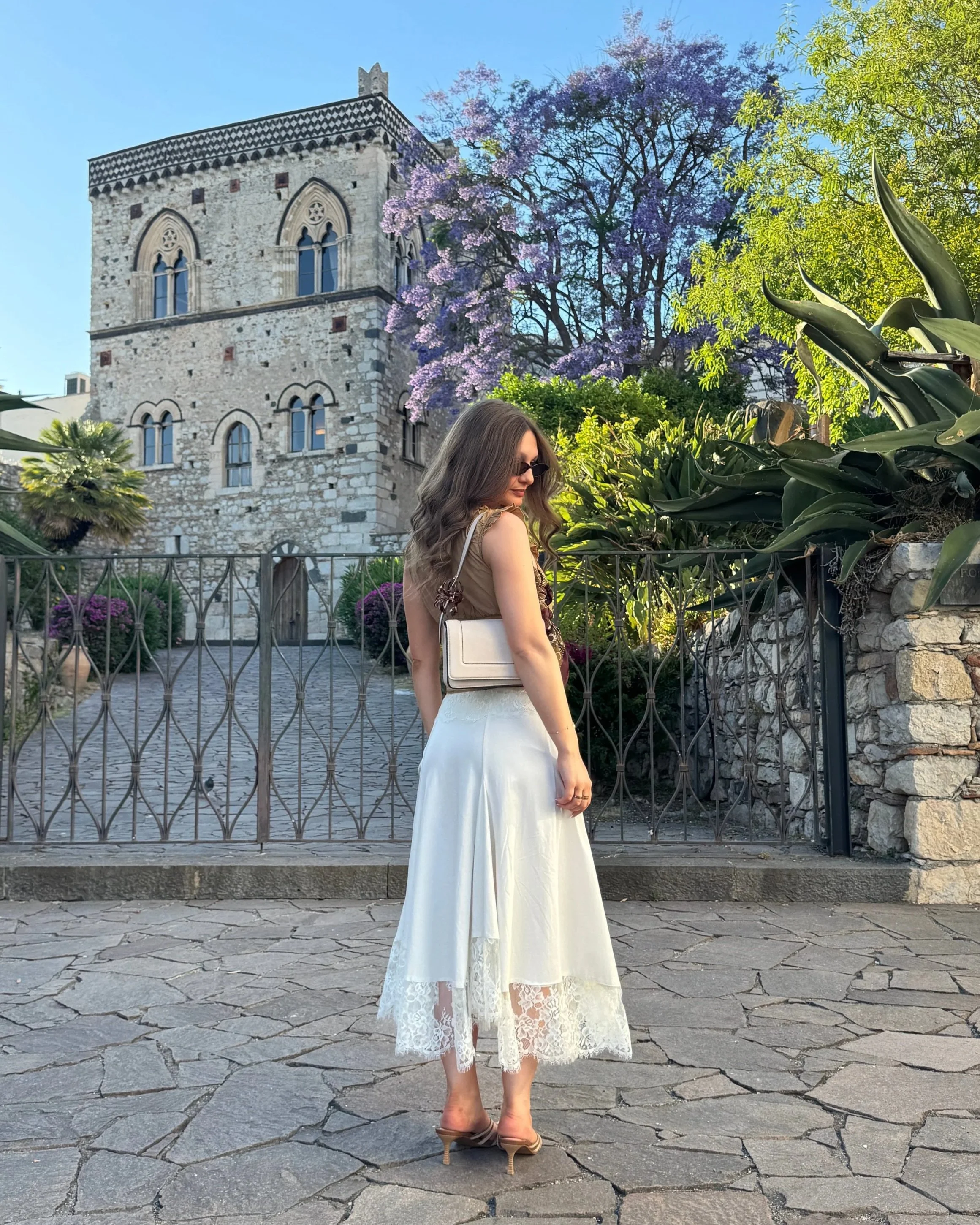 A woman in a flowing white dress stands gracefully in front of a majestic castle, surrounded by lush greenery.