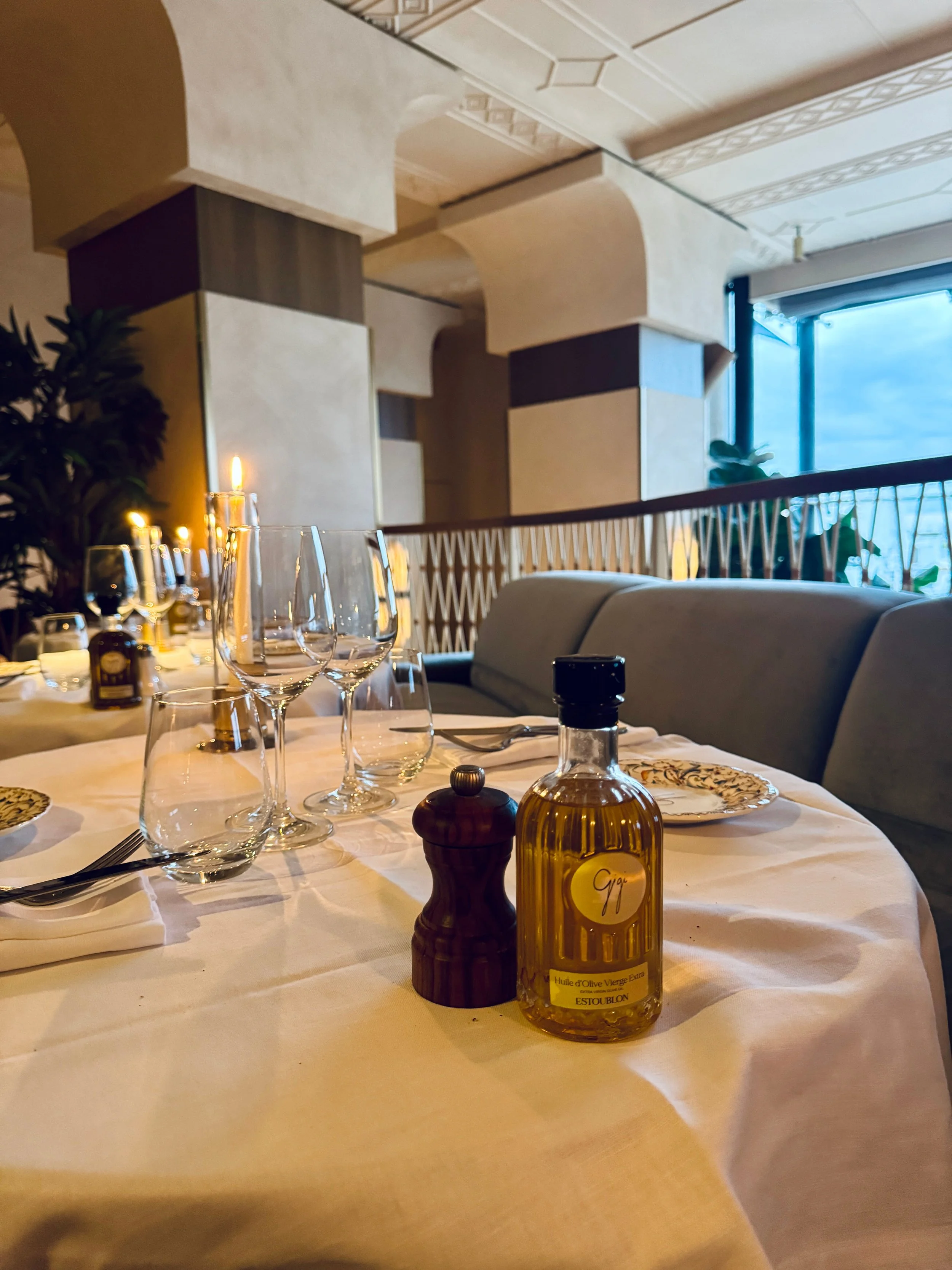 An elegant restaurant table set with wine glasses, a pepper mill, olive oil, and candlelight beside a curved booth and large windows.