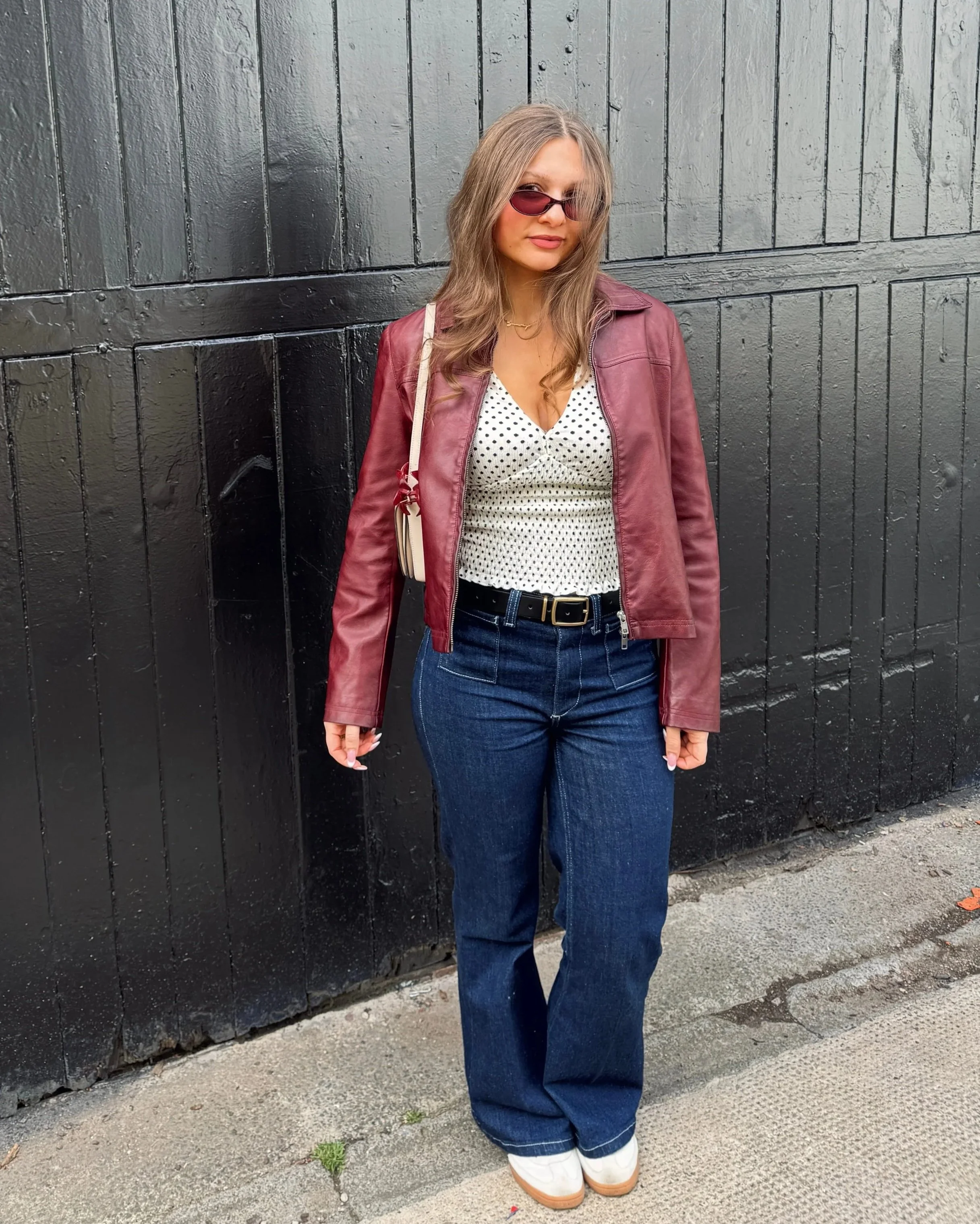 Woman in sunglasses, maroon jacket, and polka-dot top stands confidently against a black wooden background.