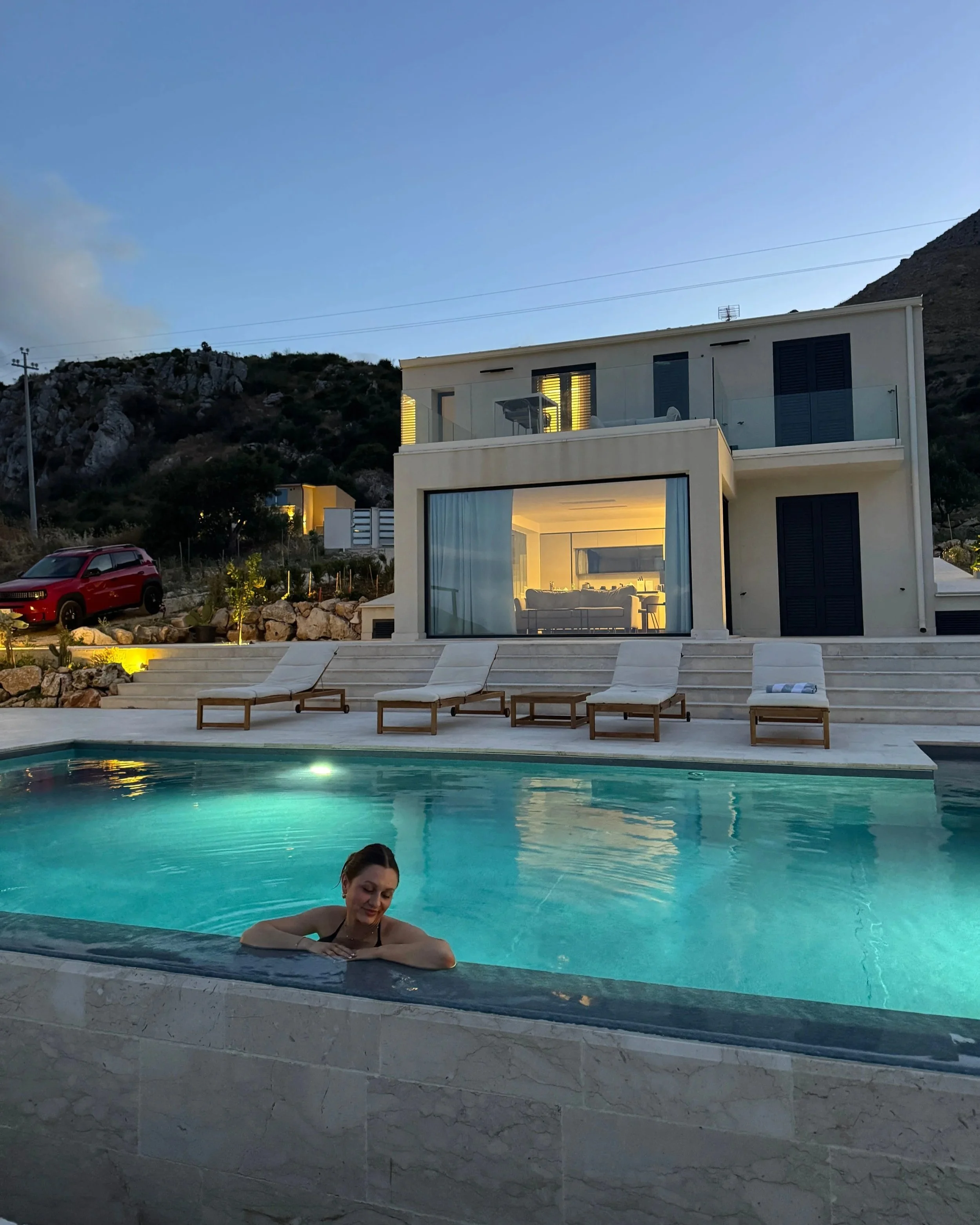 A woman sits in a tranquil pool next to a stylish home, in the evening dusk.