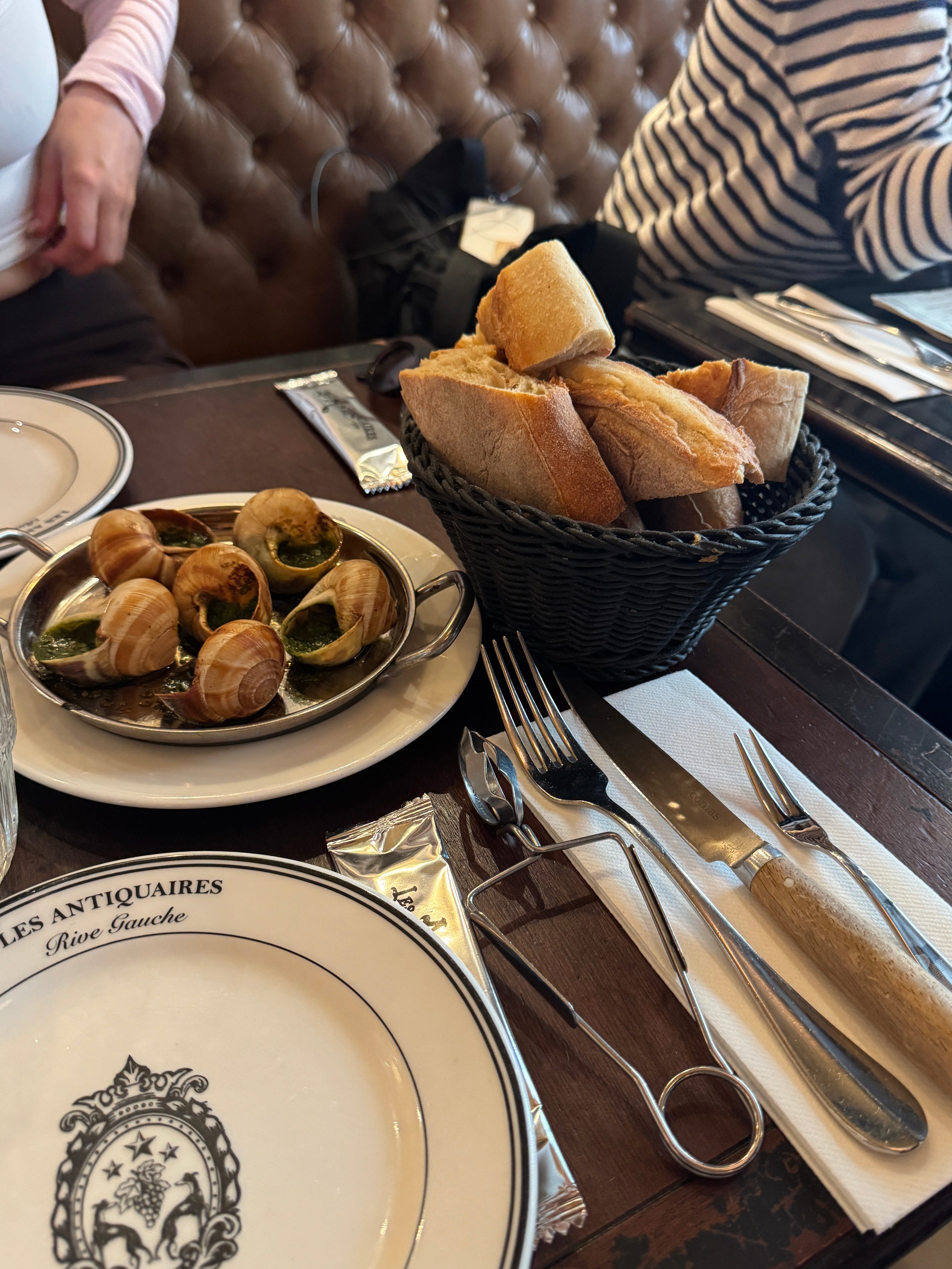 A restaurant table holds a basket of crusty bread, escargot in garlic butter, and polished cutlery as diners in striped shirts reach across; cozy, elegant dining.