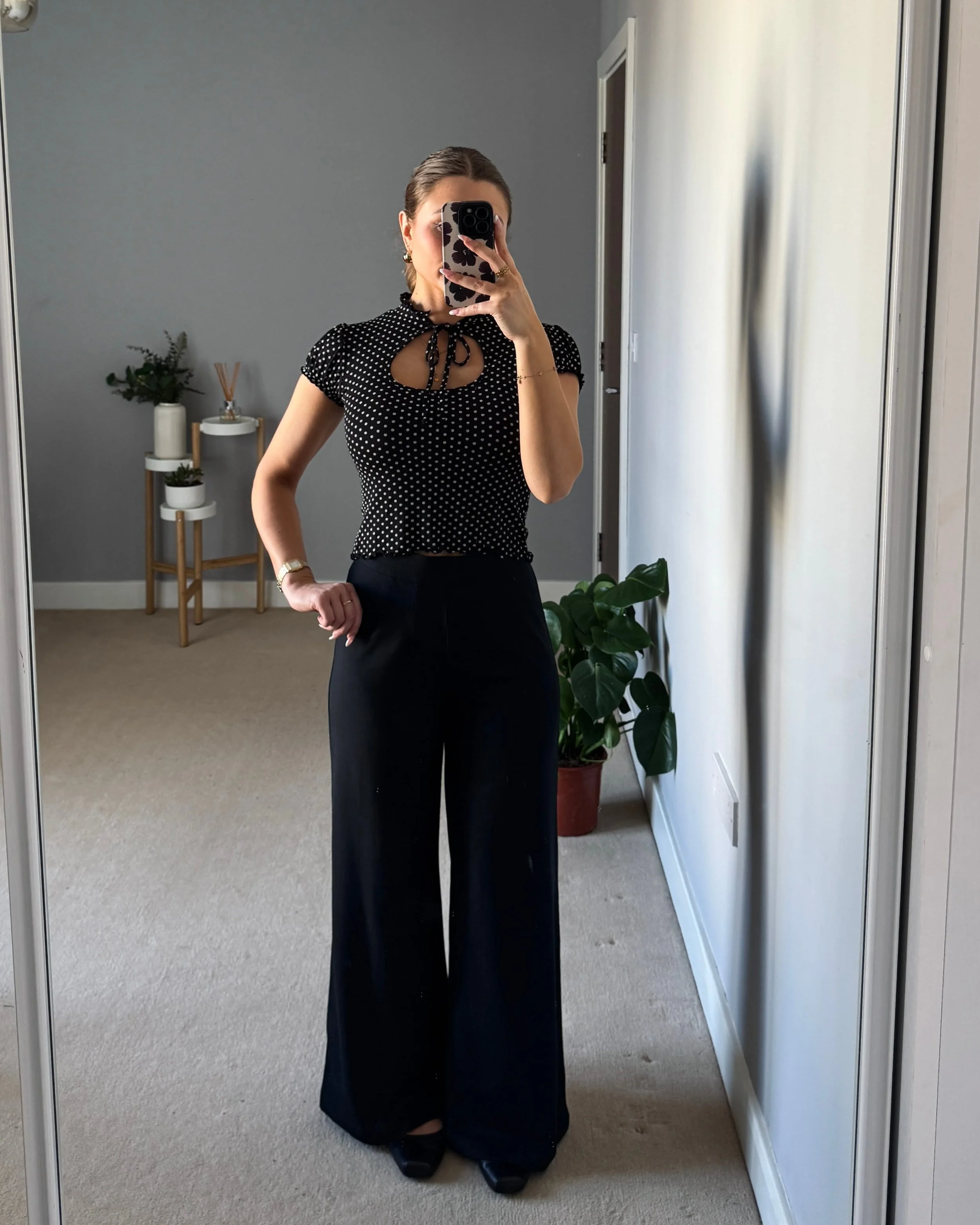 A person takes a mirror selfie in a minimalist room, wearing a black polka dot top and wide-leg pants. The background has plants and neutral decor.