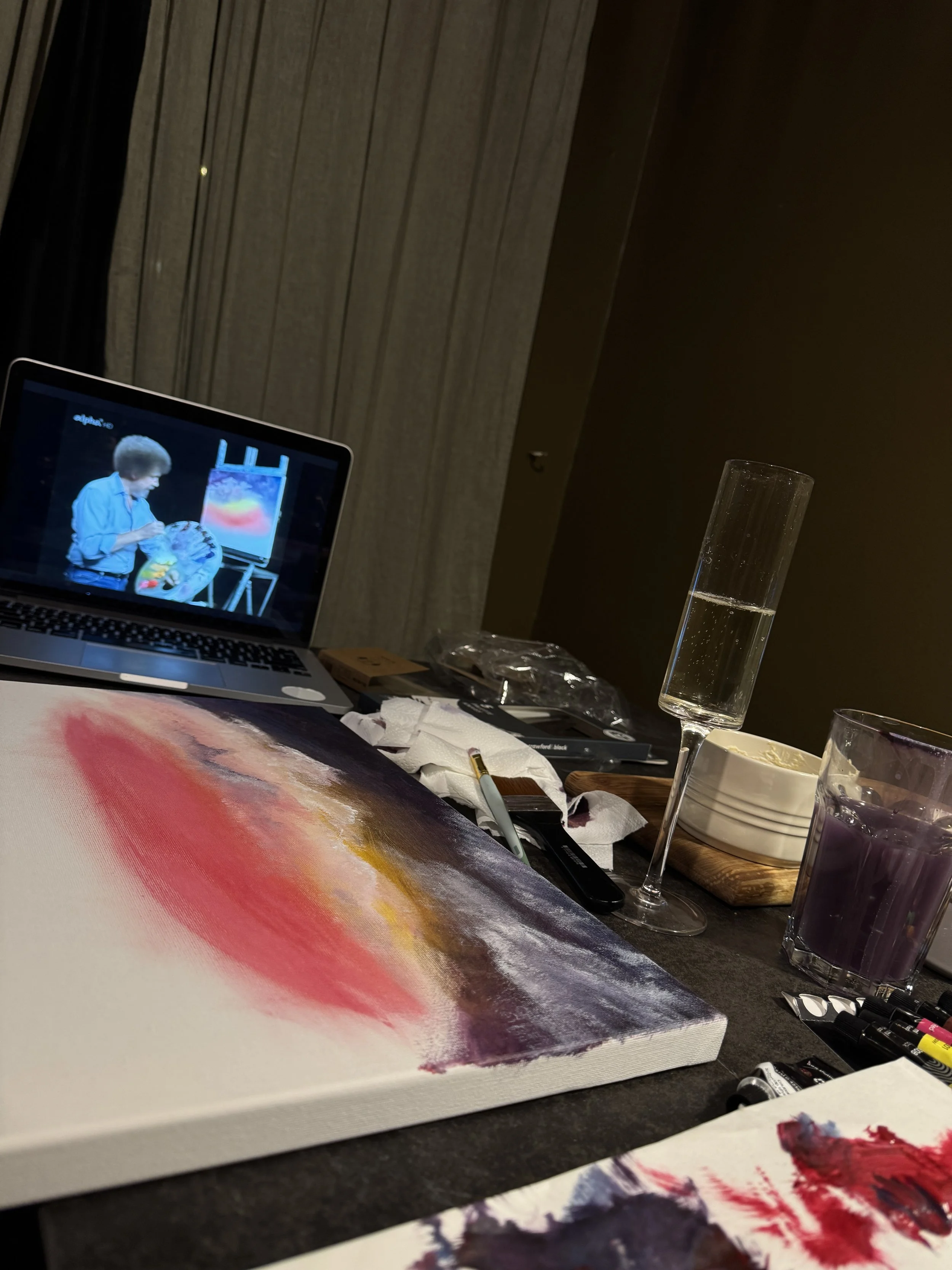 A prosecco flute, painting supplies, and a laptop playing a Bob Ross painting tutorial on a table