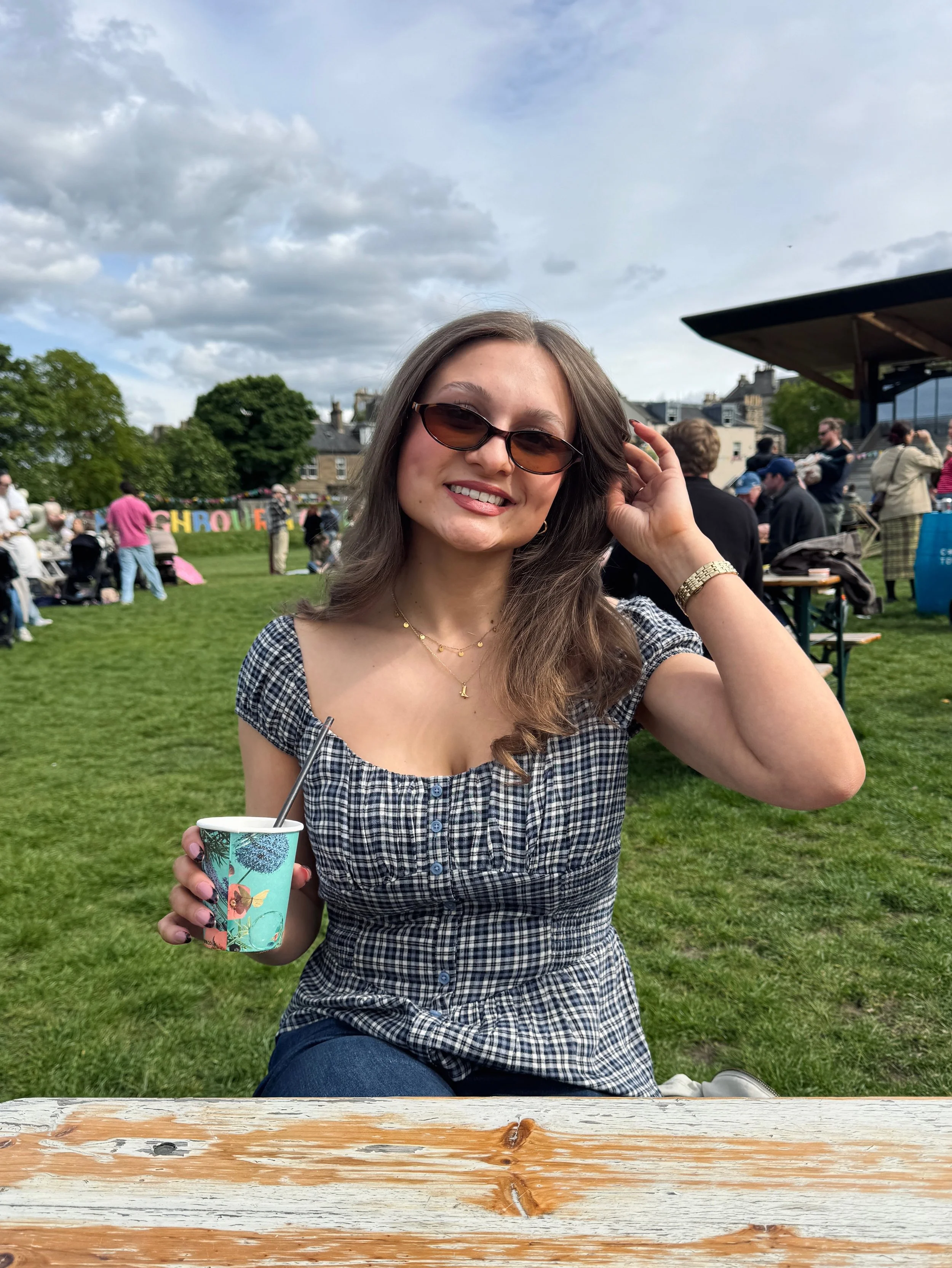 A woman in sunglasses smiles while sitting outdoors on a sunny day. She holds a colorful cup with a straw, surrounded by people on green grass.