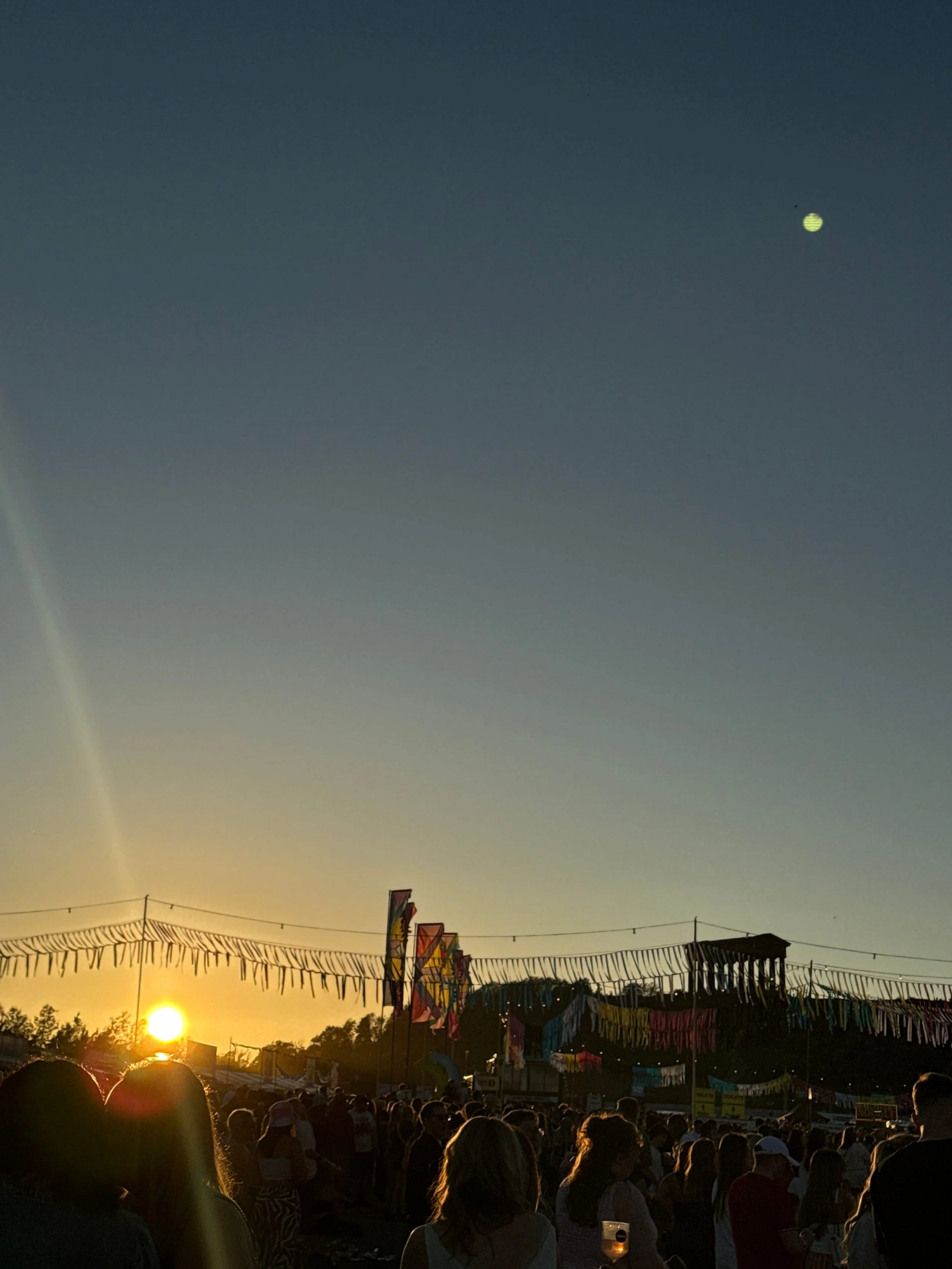 A bustling crowd at a festival during sunset, surrounded by colorful flags and a warm, inviting sky.