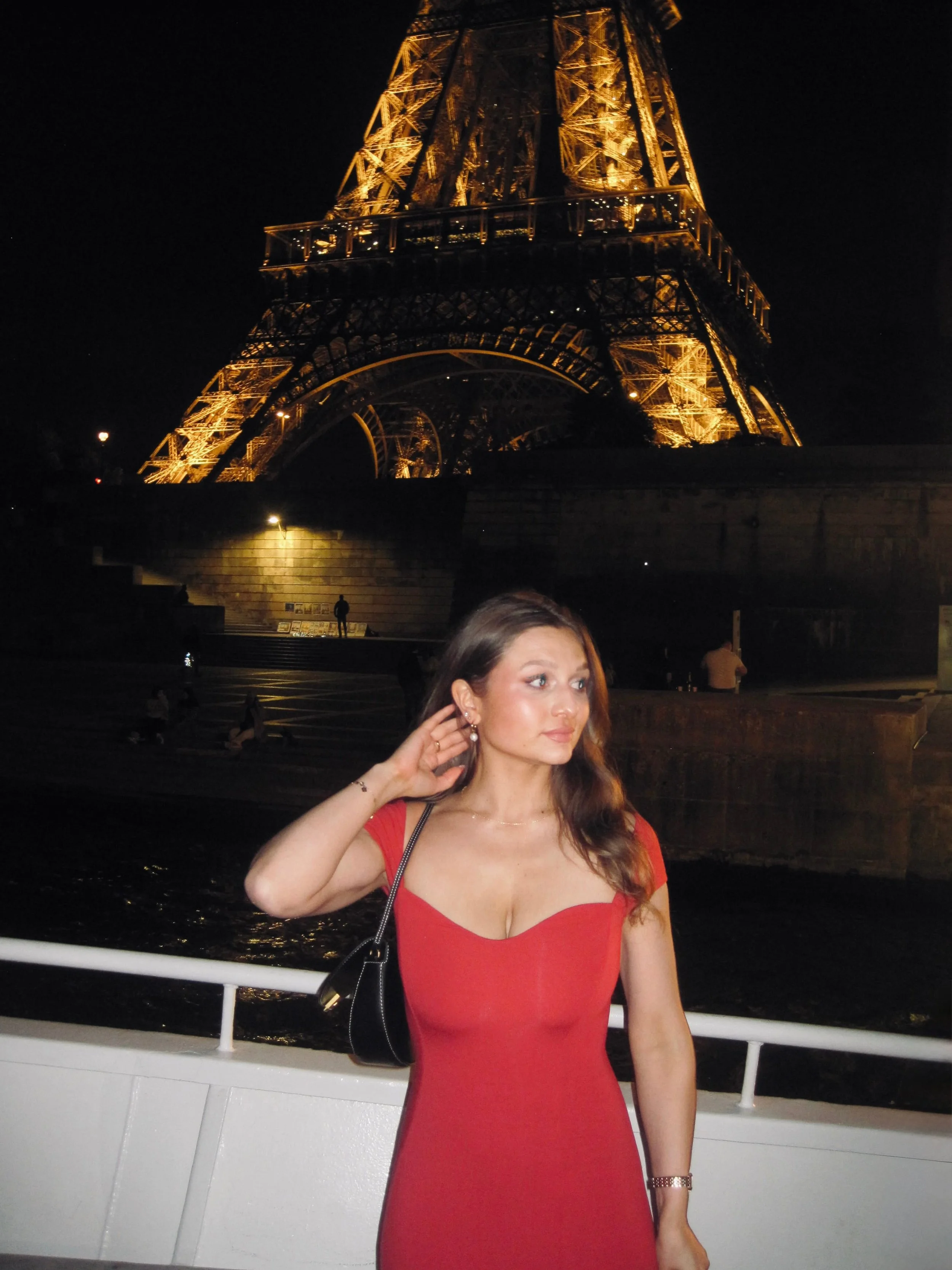 Woman in a red dress poses on a boat at night, with the warmly illuminated Eiffel Tower rising behind her in Paris, creating an elegant mood.