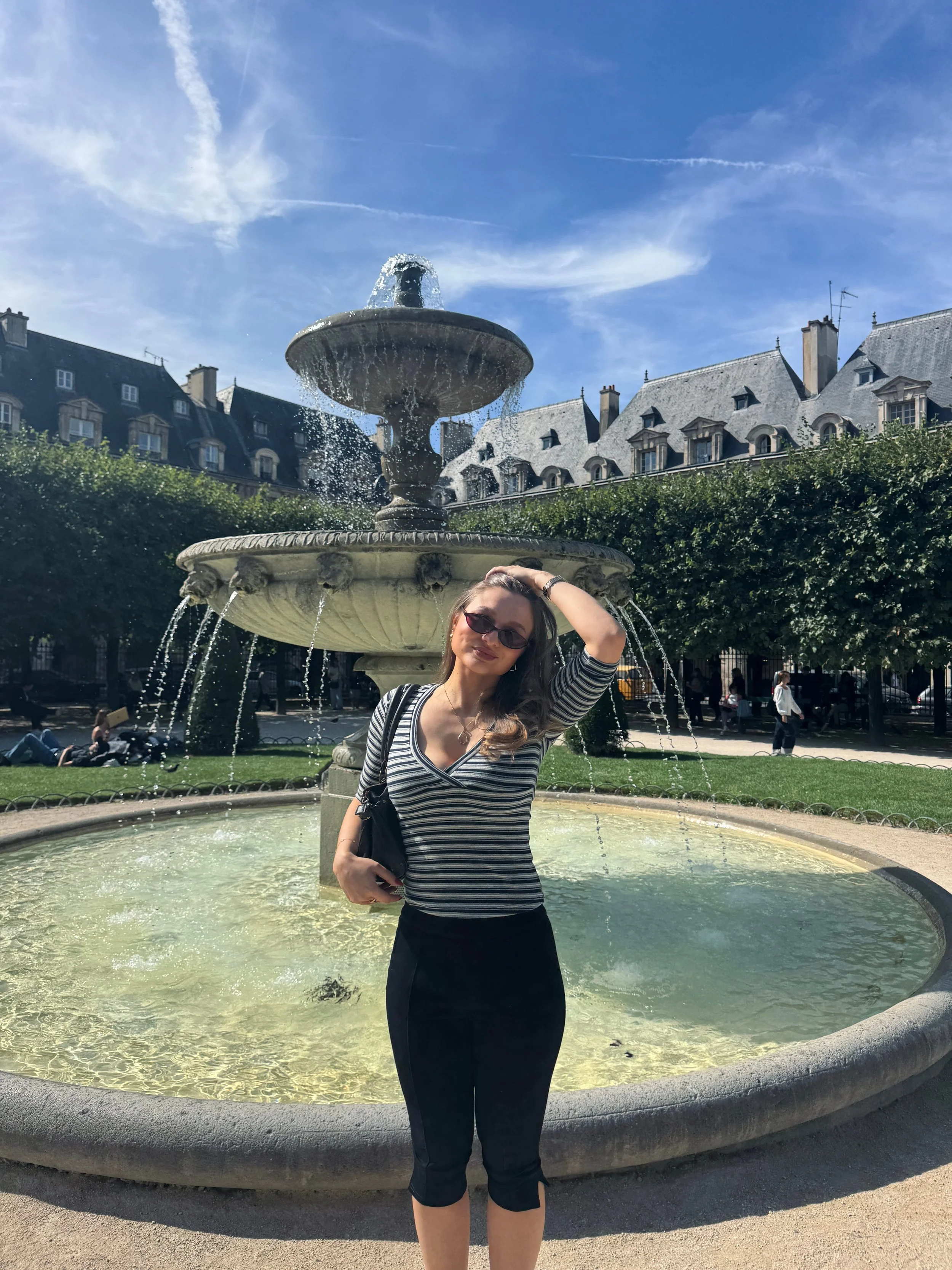 Woman in sunglasses poses beside a tiered fountain in a sunny plaza, with historic buildings, green hedges, and bright blue sky behind her.