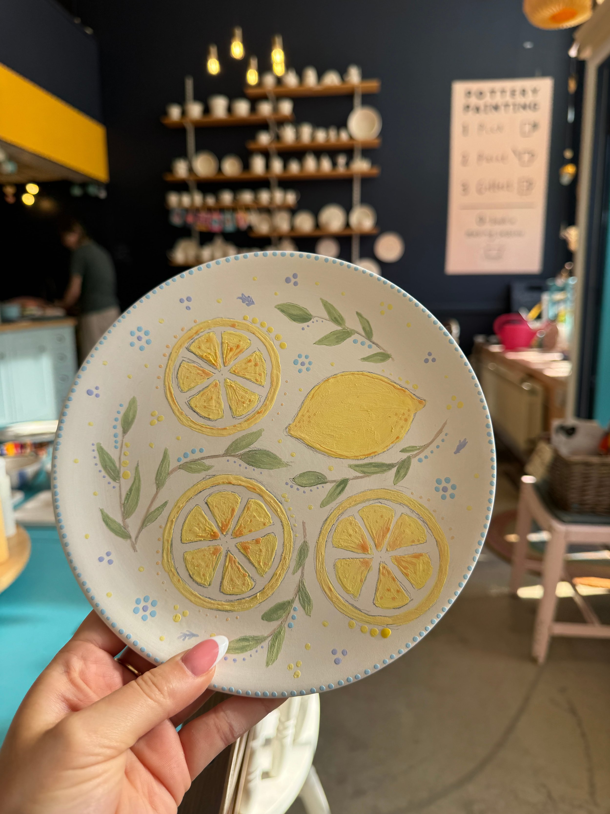 A hand holding up a painted plate with a lemon design at a pottery painting experience