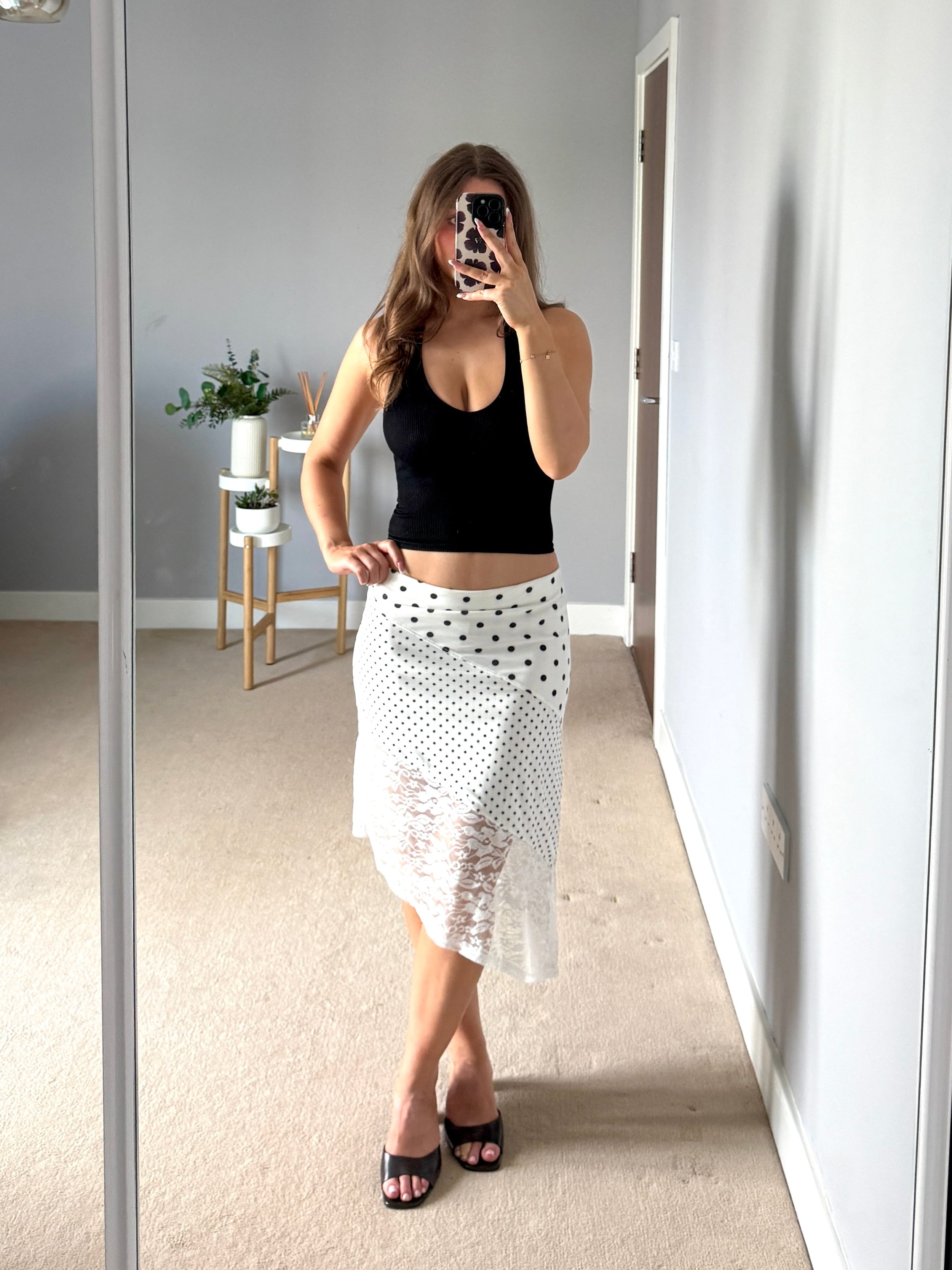 Woman taking a mirror selfie wearing a black sleeveless top, white skirt with polka dots and lace, and black open-toe heels in a minimalist room.