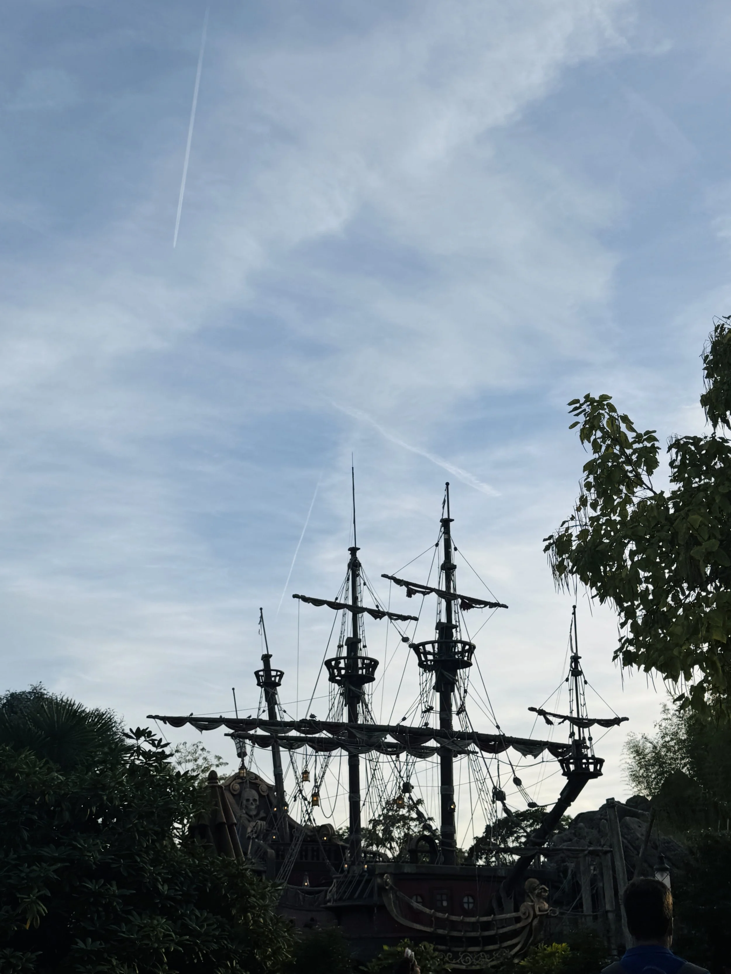 A historic wooden sailing ship with tall masts and rigging sits among trees beneath a broad, cloudy blue sky, creating a quiet, nostalgic mood.