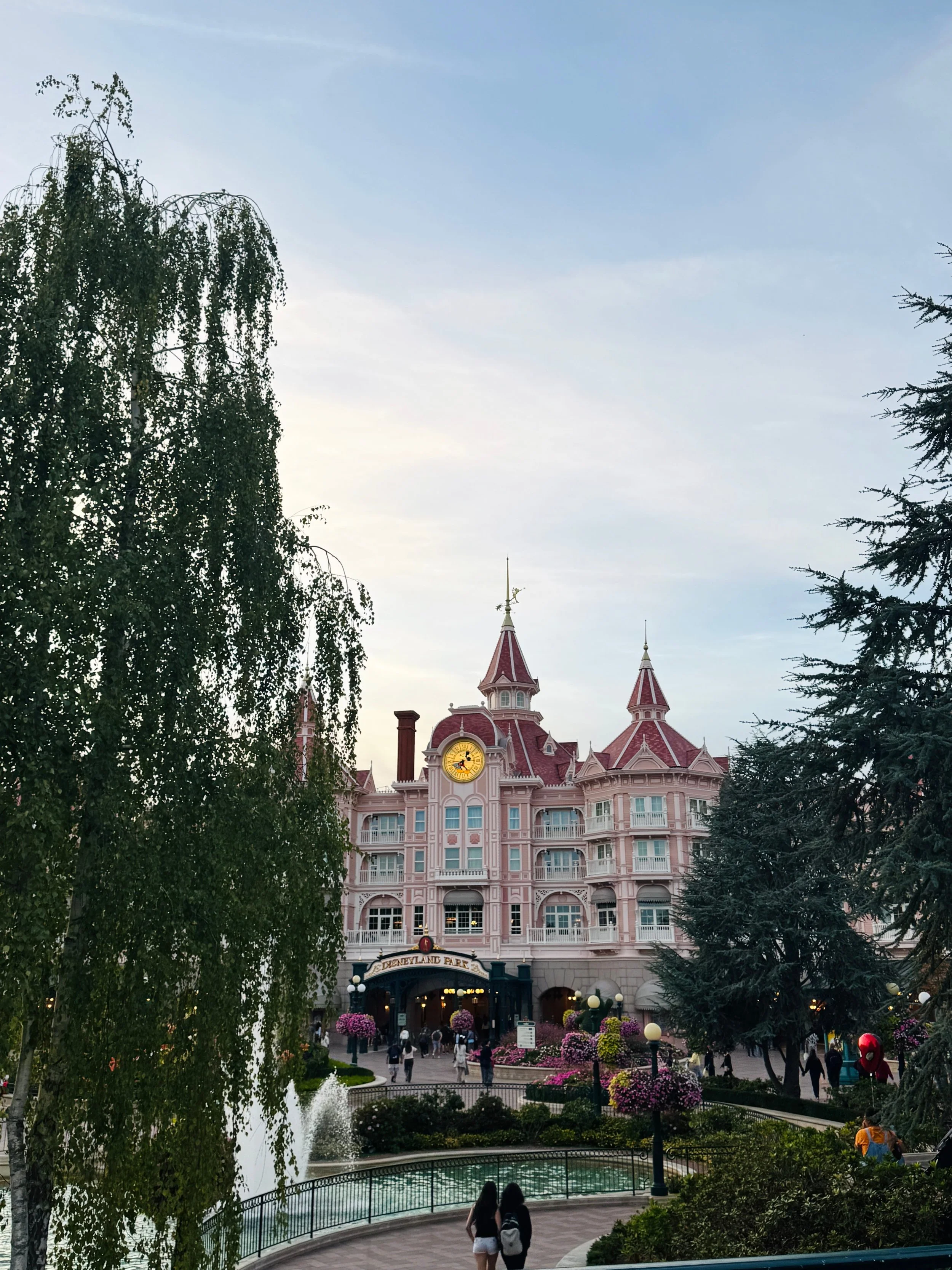 A pink, turreted Victorian-style hotel stands beyond a garden pond, framed by tall trees and flowers as visitors stroll beneath a calm, cloudy sky.