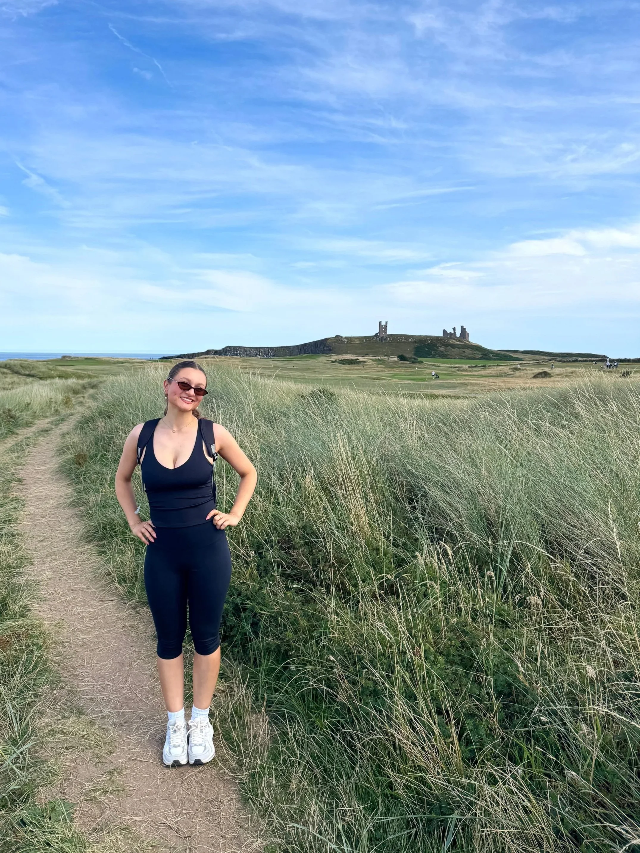Smiling woman in sunglasses poses on a sandy path through coastal grass, with sea, castle ruins, and a bright blue sky behind her.
