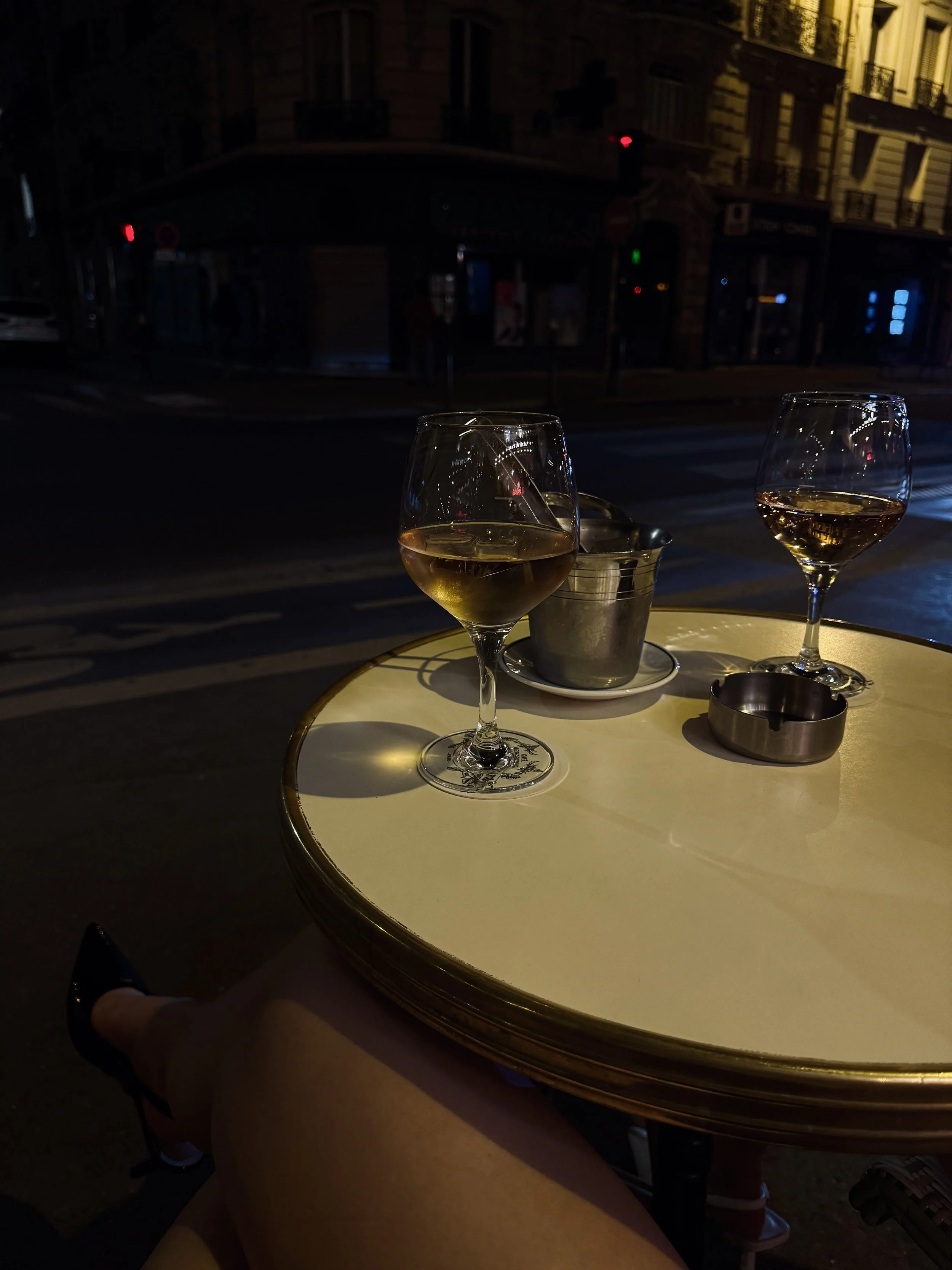 At a dim nighttime sidewalk café, two glasses of amber wine sit on a round cream table beside an ice bucket and ashtray, with a quiet city street beyond.
