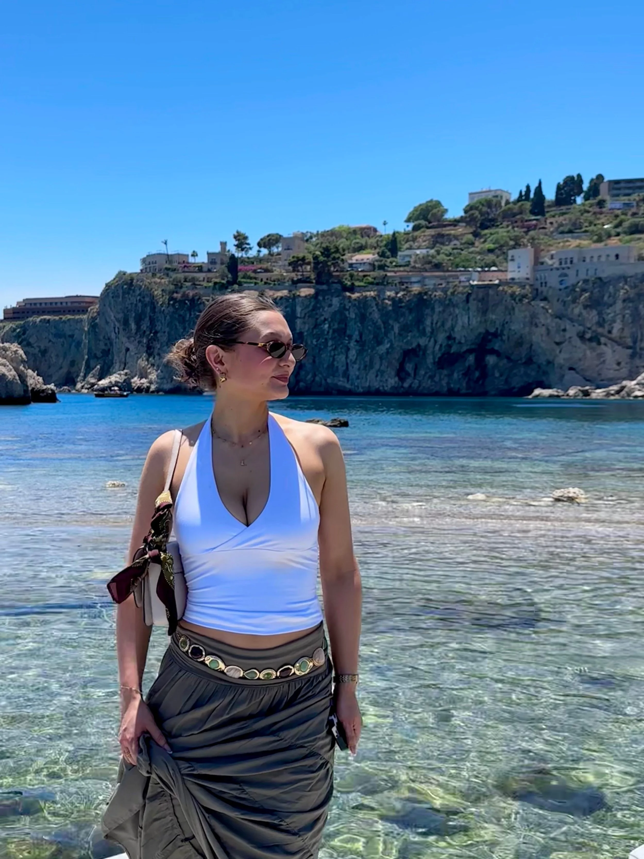 Woman in white halter top and long skirt standing in clear shallow water with rocky cliffs and buildings in the background under a blue sky.