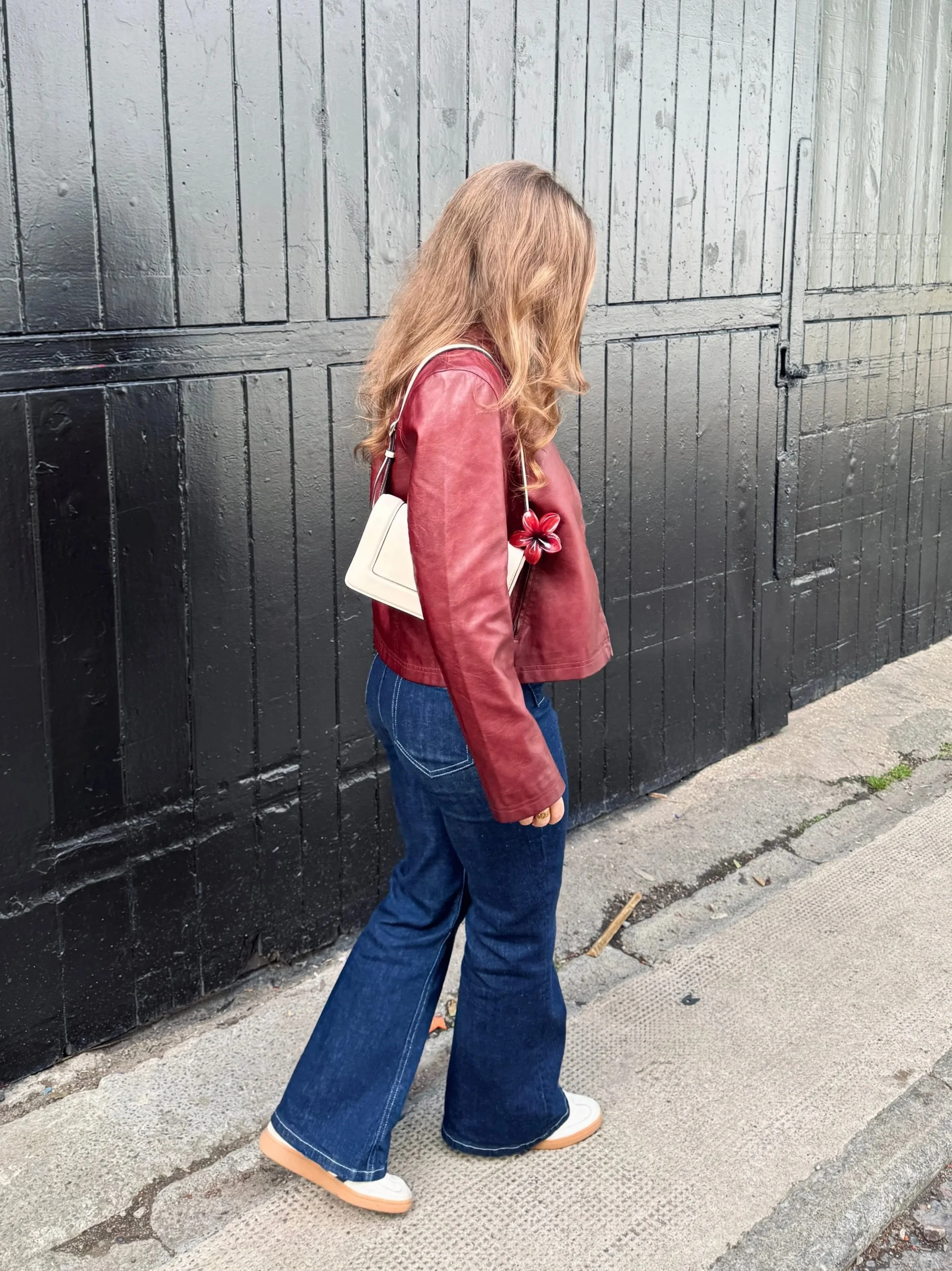 A woman in a red leather jacket and blue jeans walks along a textured sidewalk. She carries a white handbag, with her hair cascading down. The mood feels casual and relaxed against a large black wooden gate backdrop.