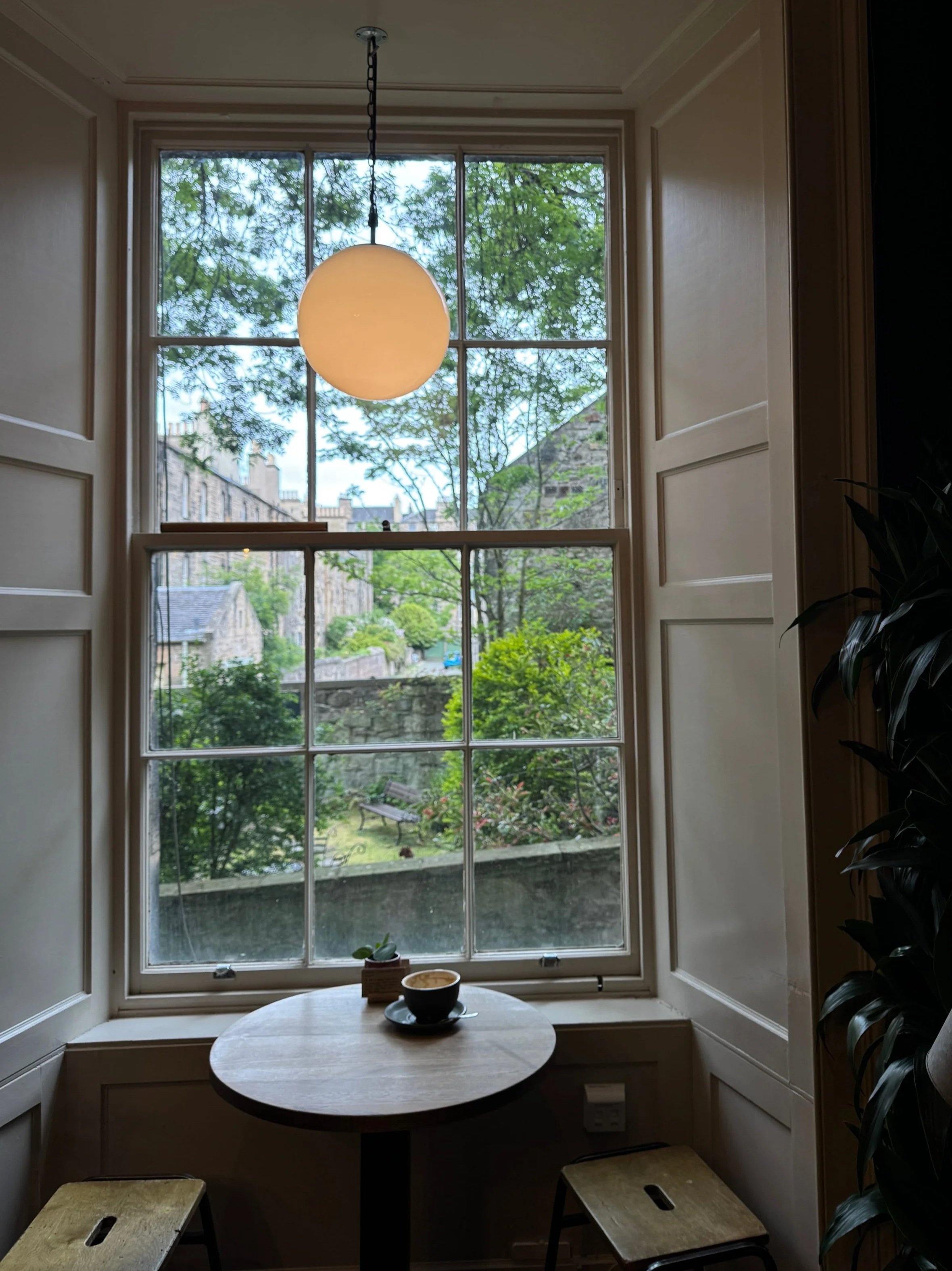 Cozy cafe nook with a round table, a cup of coffee, and two stools. Large window view of lush garden and historic buildings, soft pendant light above.