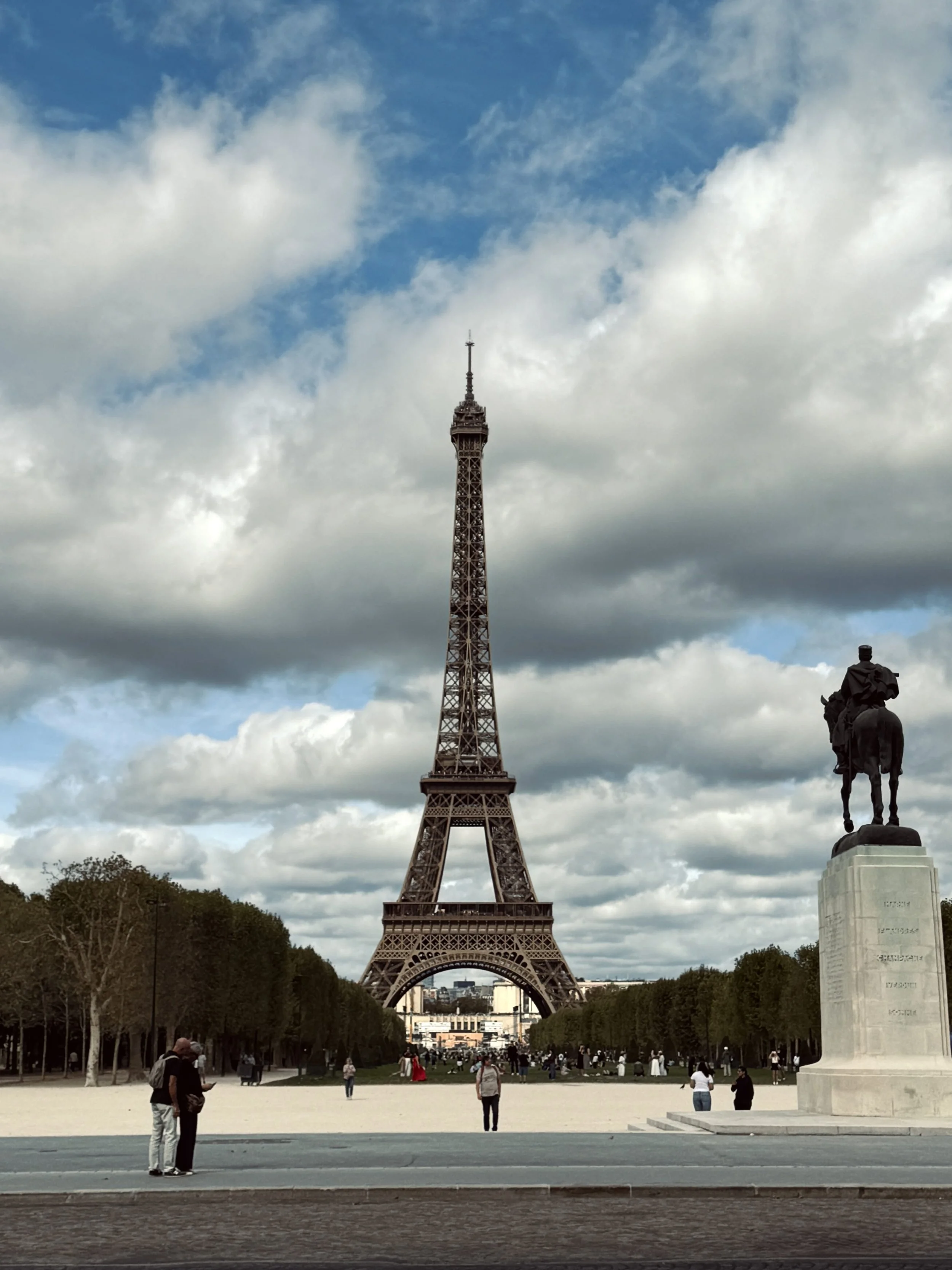 The Eiffel Tower rises over a broad Paris plaza beneath dramatic clouds, with a statue, trees, and scattered visitors creating a grand, open atmosphere.